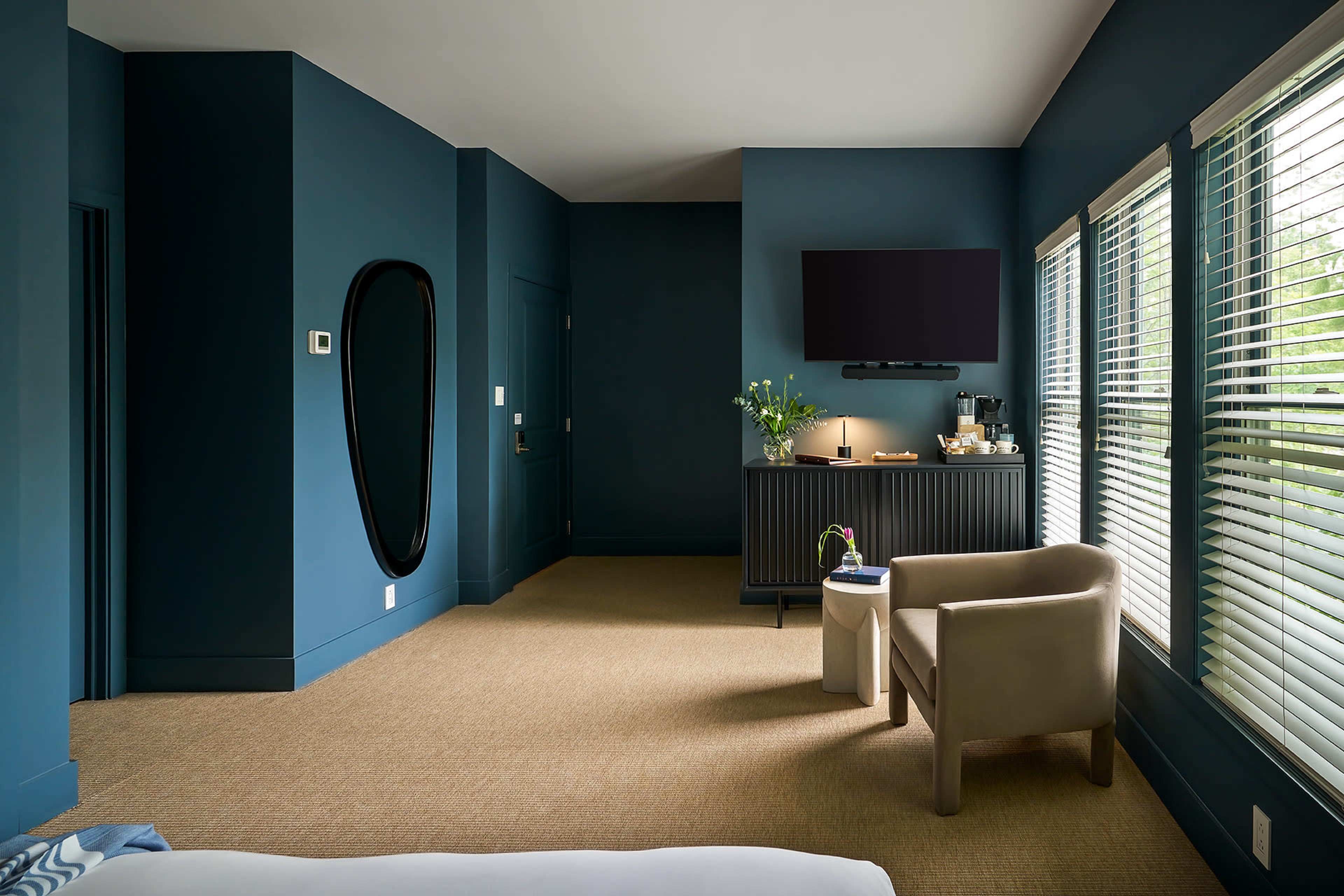 A modern, well-lit hotel room featuring a blue color scheme, a large wall mirror, a flat-screen TV, and a small seating area with a chair and side table.