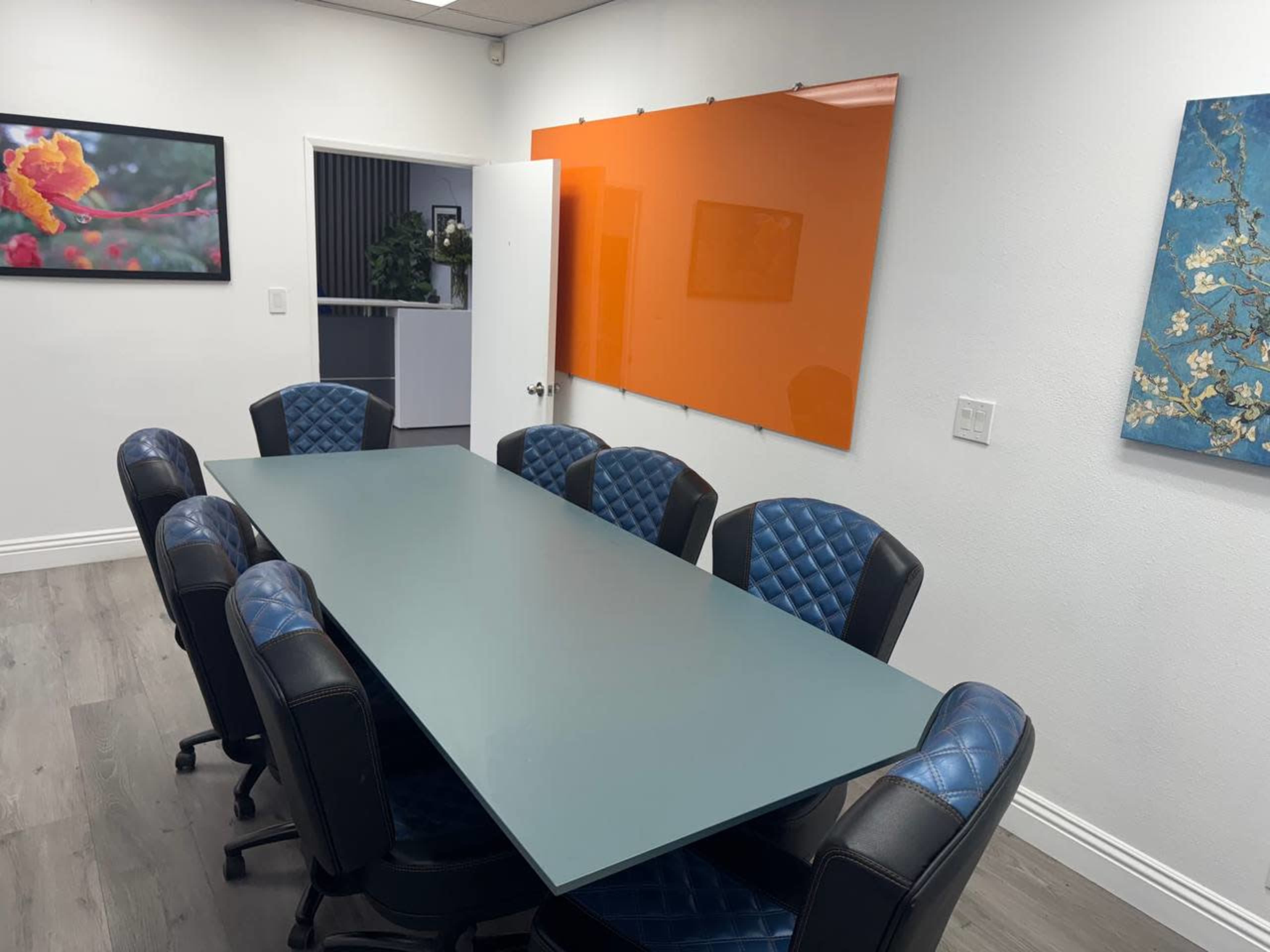 The image shows a modern conference room featuring a long rectangular table surrounded by eight upholstered chairs, with an orange board and decorative wall art in the background.
