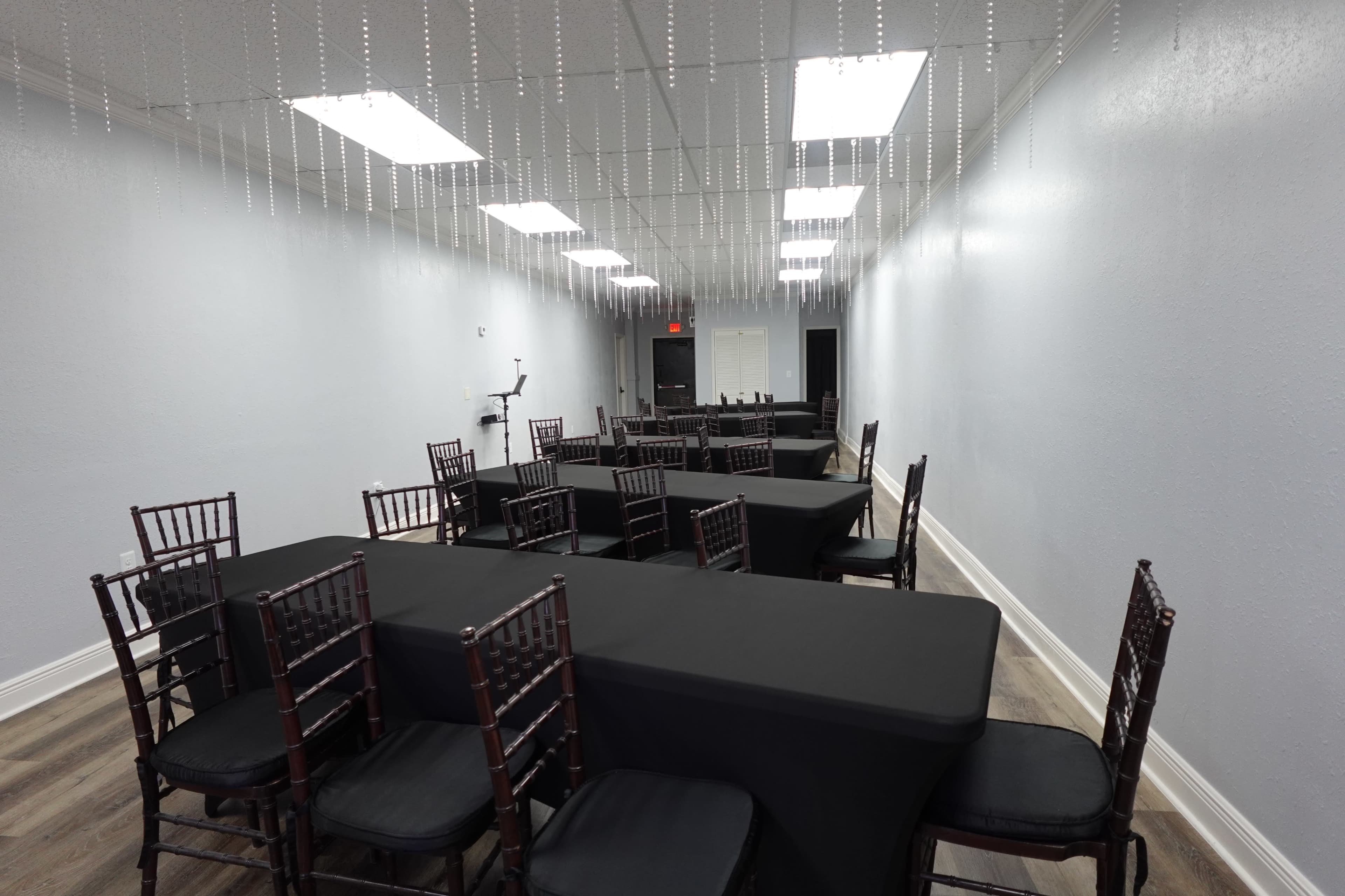 A long, narrow event room features black tables and chairs arranged in a formal setup under a suspended decorative element.