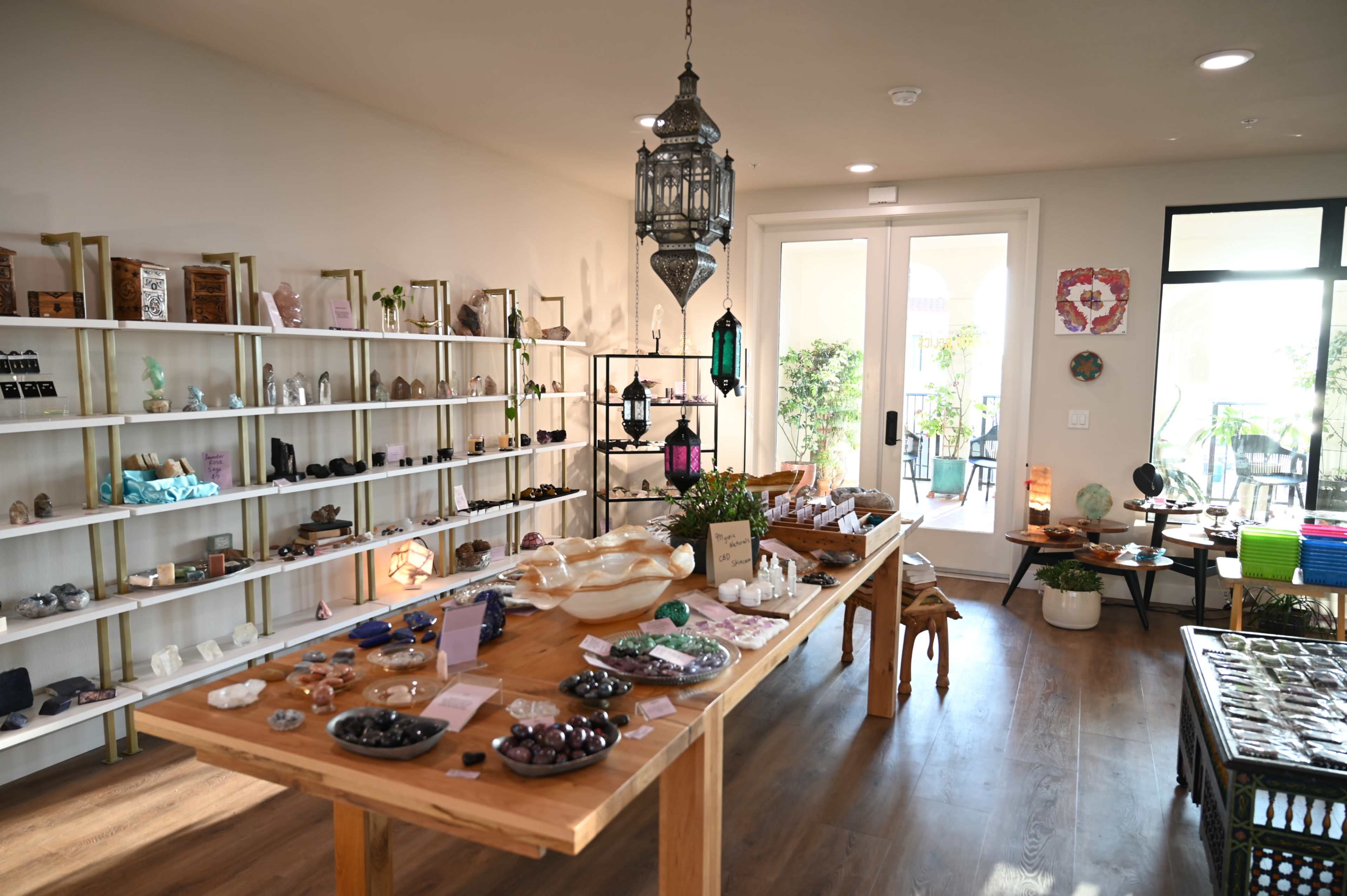 The image shows a well-organized shop interior featuring wooden tables displaying various crystals and decorative items, with shelves filled with similar merchandise along the walls.