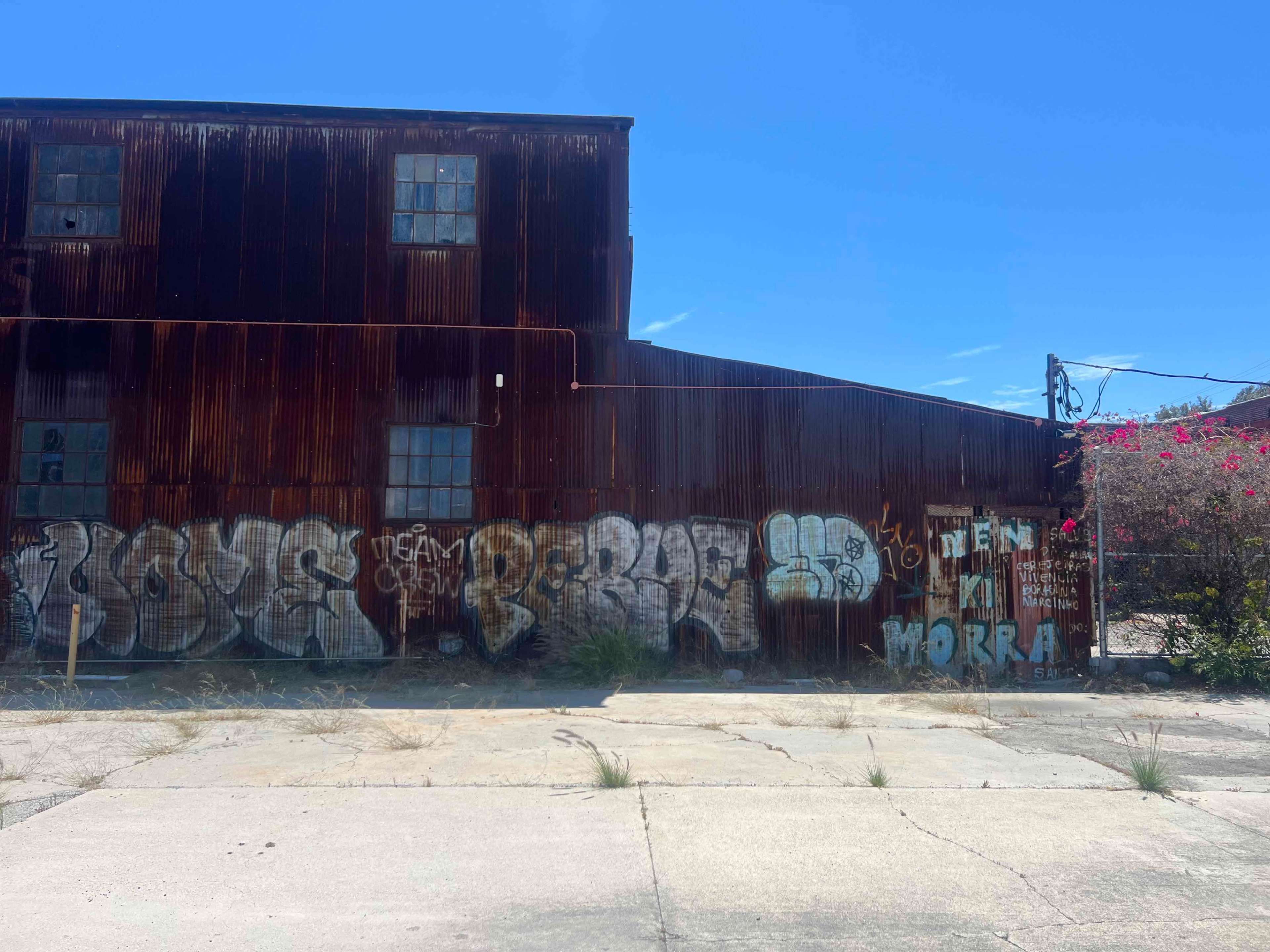 A weathered industrial building features rusted metal siding and graffiti-covered walls, with patches of overgrown grass on the concrete below.