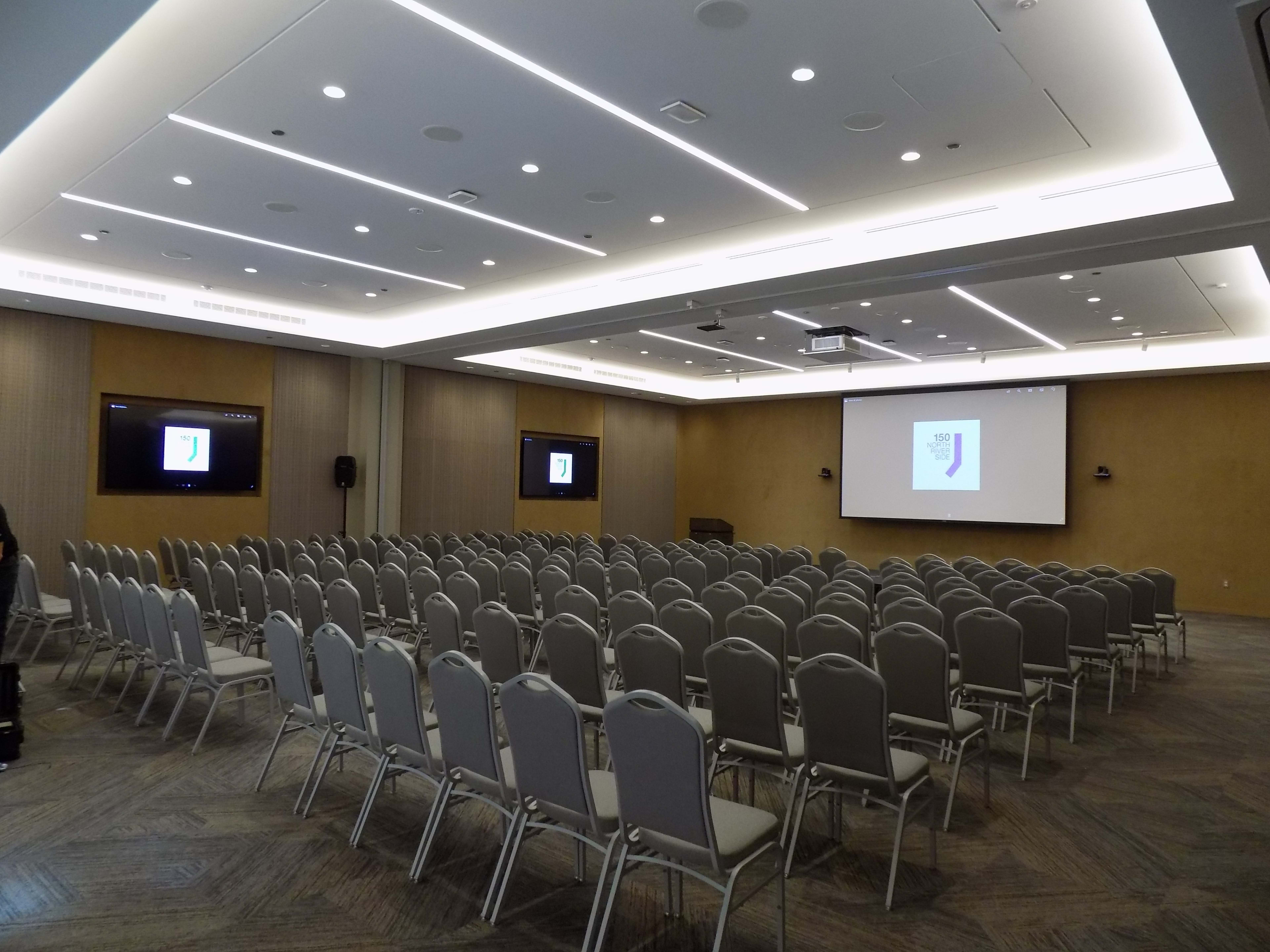 A conference room is set up with rows of empty chairs facing two screens displaying a logo.