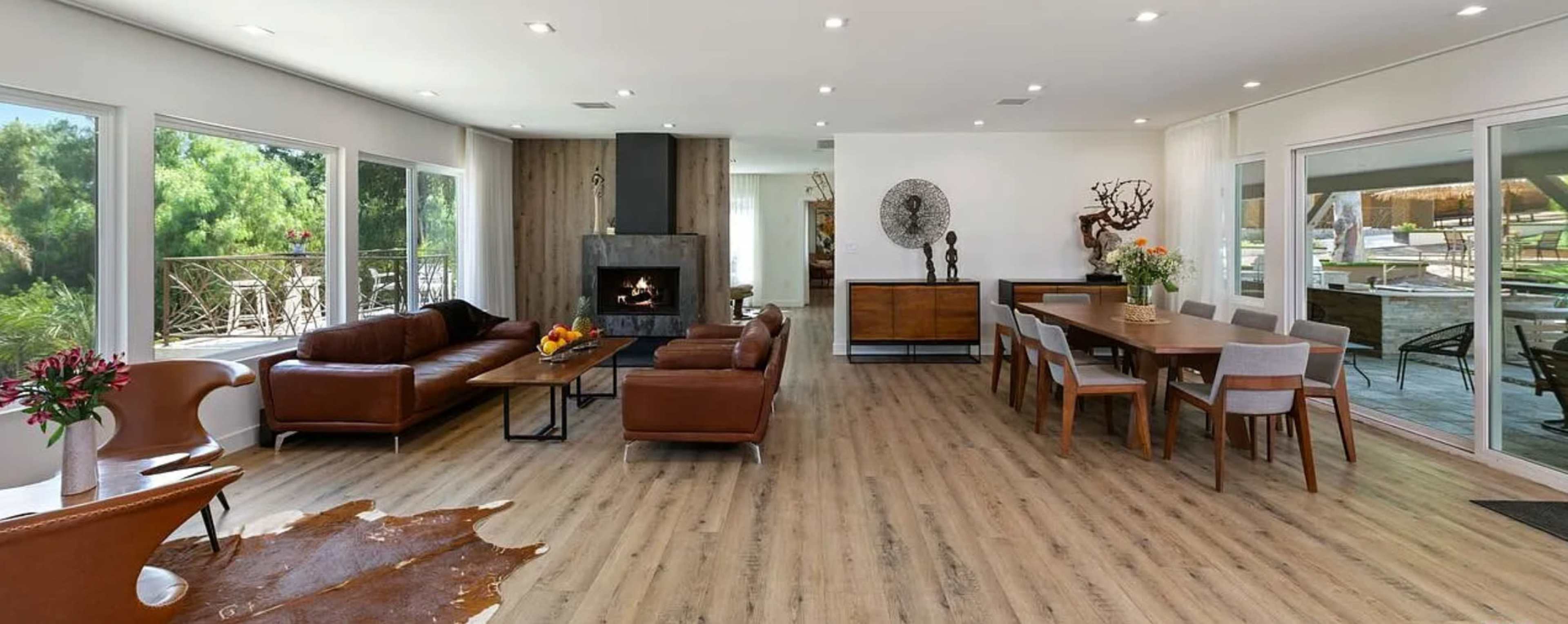 The image depicts a modern living room with a fireplace, a large dining table, and large windows that provide ample natural light.