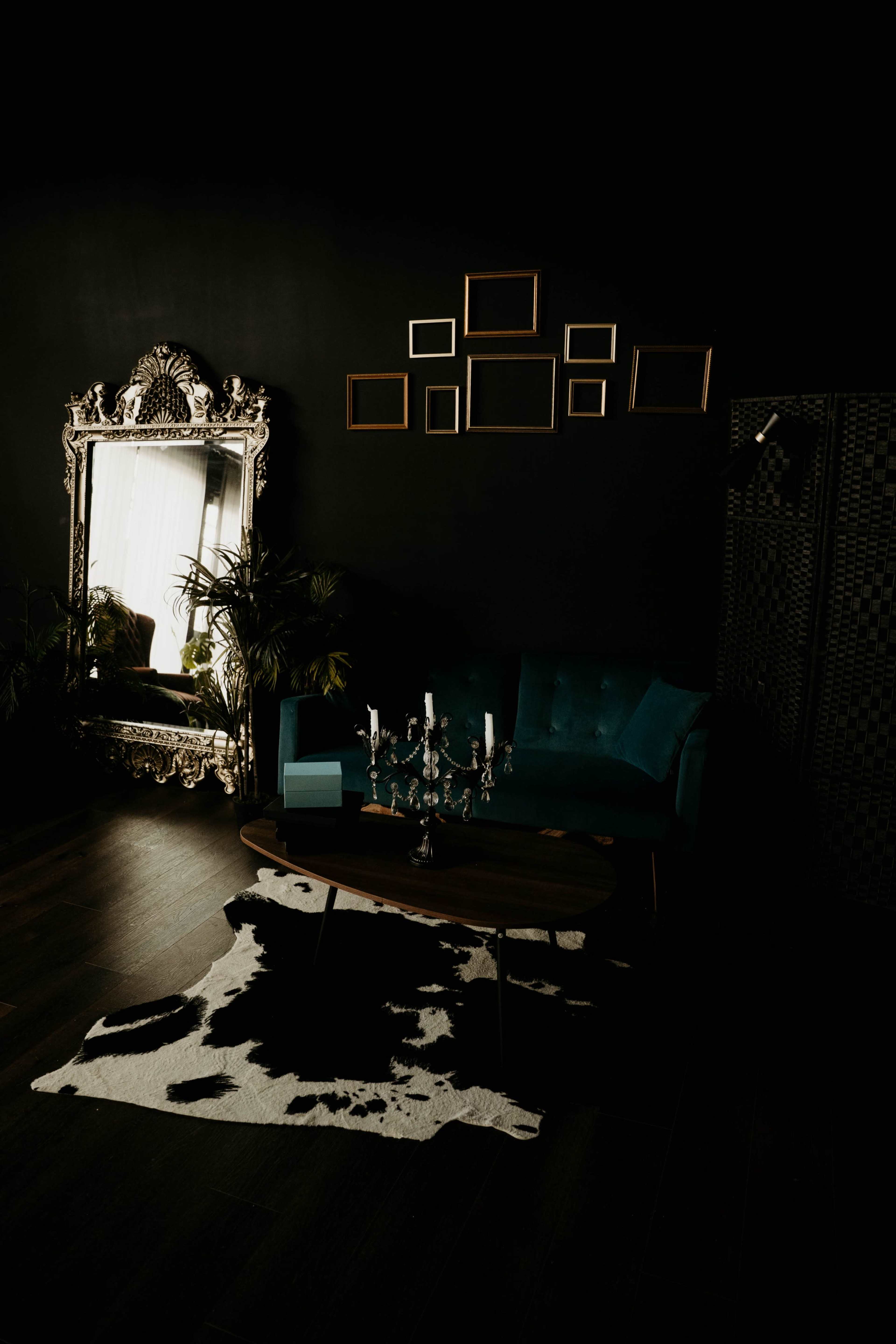 The image depicts a dimly lit living room with a dark wall, a teal couch, a large ornate mirror, and a coffee table with a candle holder, placed on a cowhide rug.