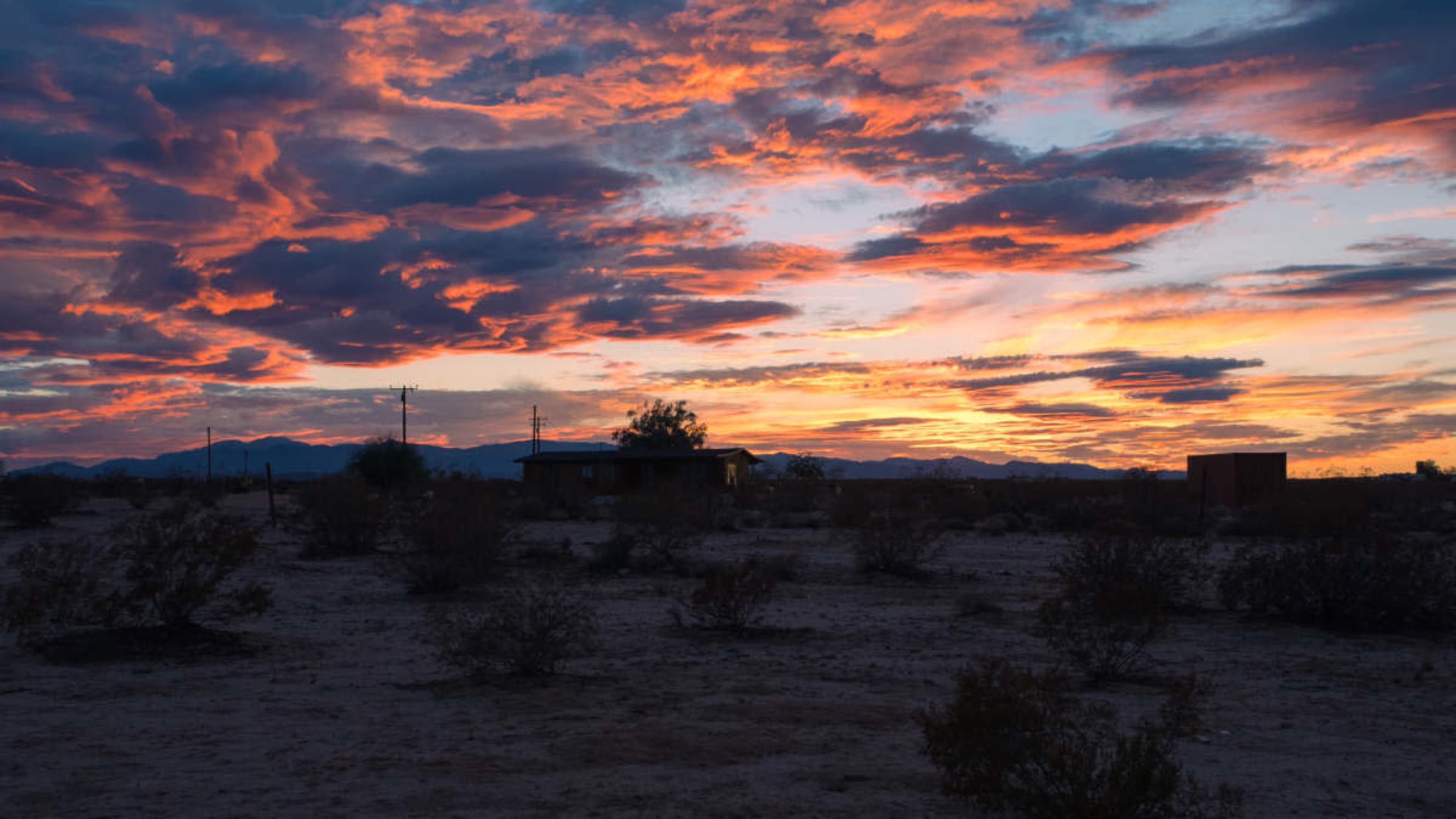 A colorful sunset illuminates the sky over a desert landscape with sparse vegetation and a distant silhouette of mountains.