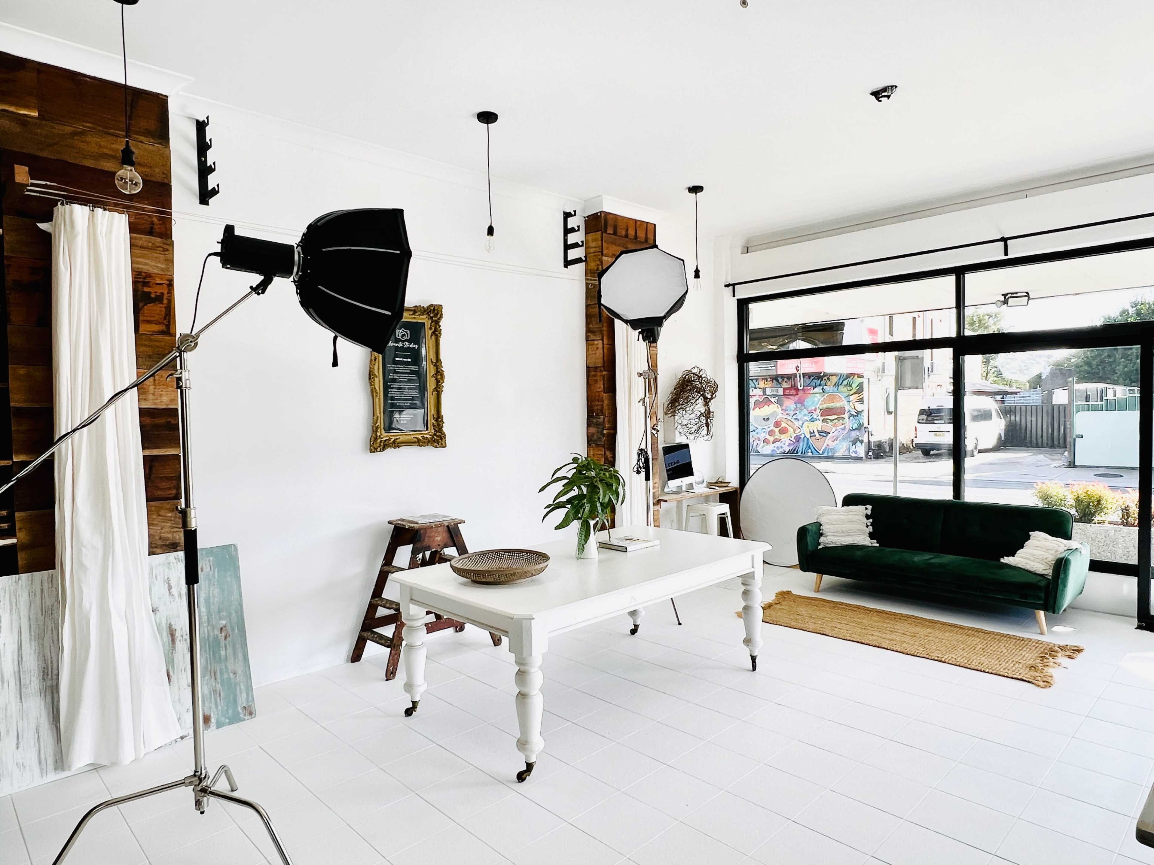 The image shows a minimalist studio space featuring a large table, two lighting setups, a green sofa, and a window displaying a colorful mural outside.