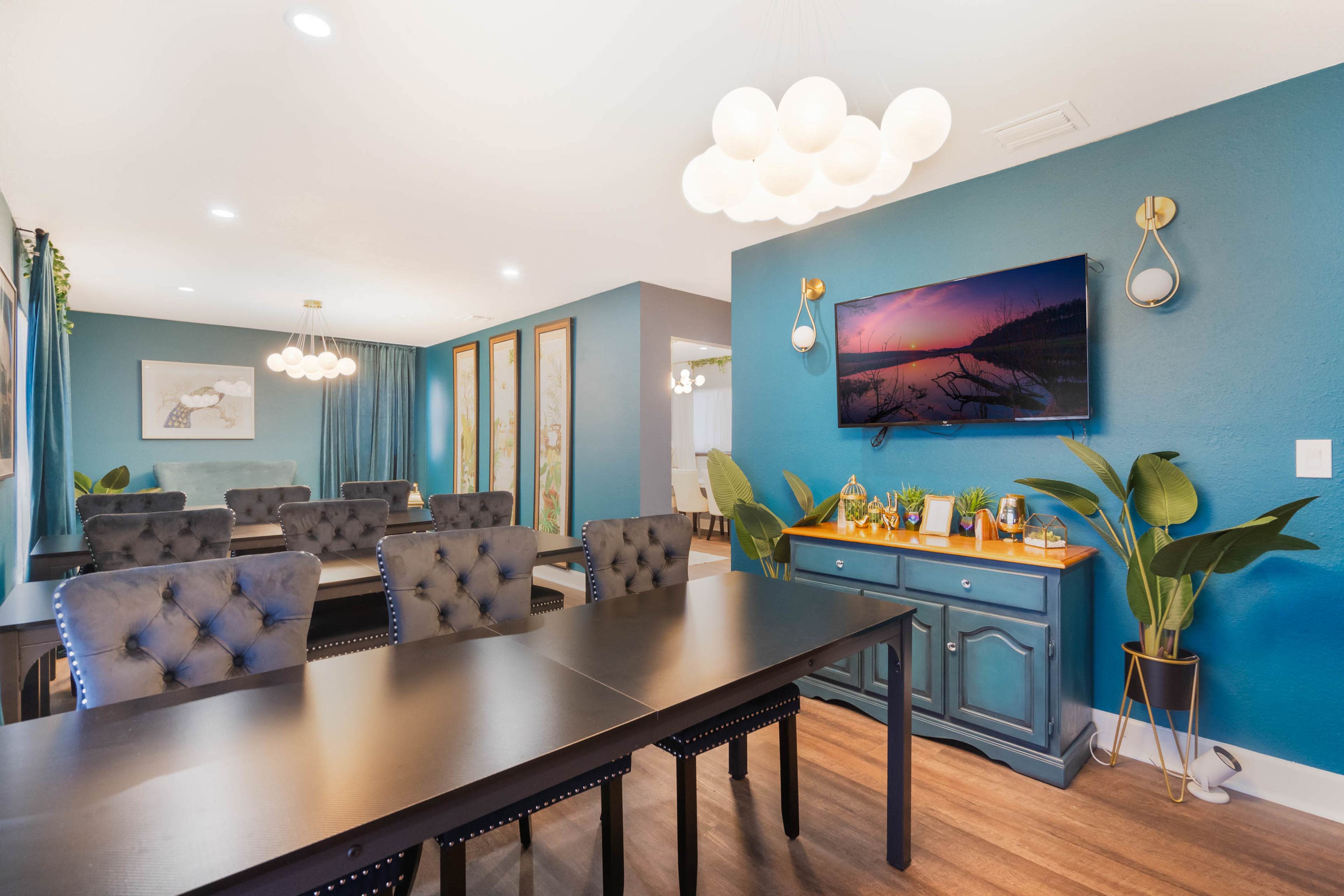The image shows a modern meeting room featuring dark tables and upholstered chairs, with a wall-mounted television and indoor plants framed by teal-colored walls.