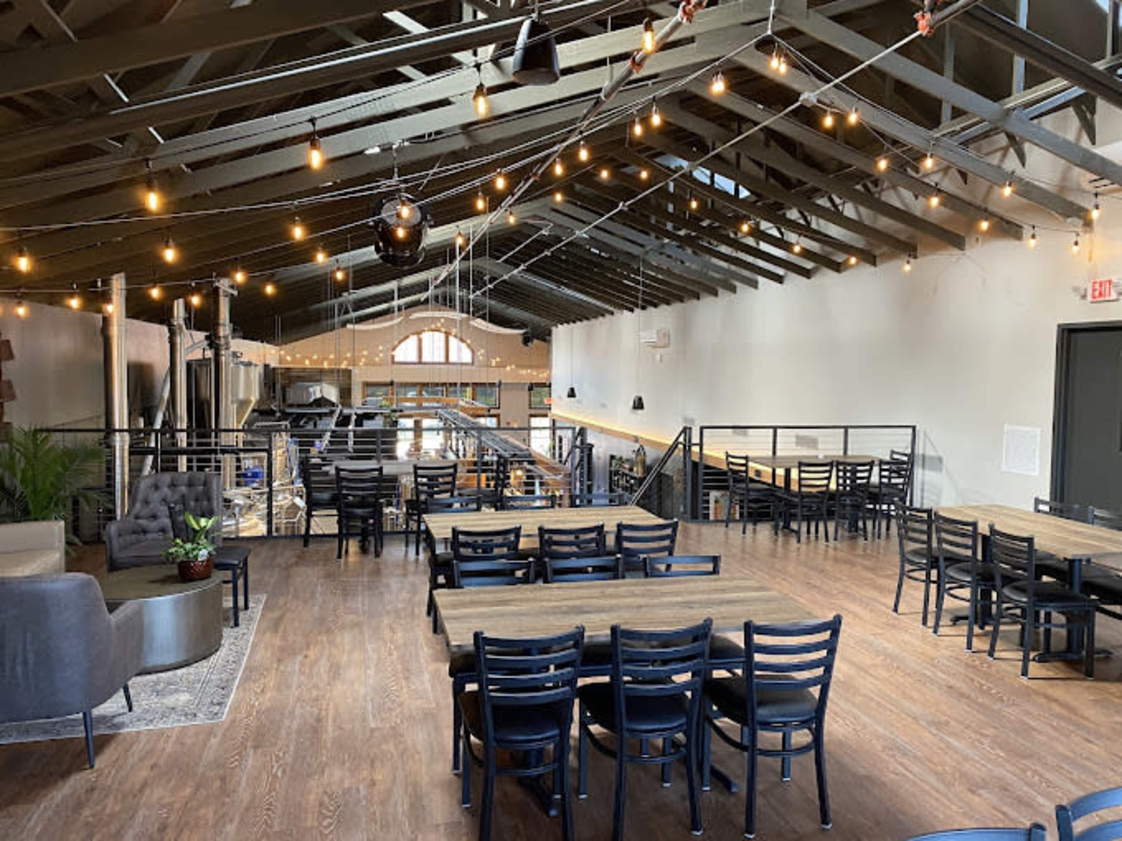 The interior of a modern, open-concept restaurant features wooden floors, multiple dining tables, and string lights hanging from the high ceiling.