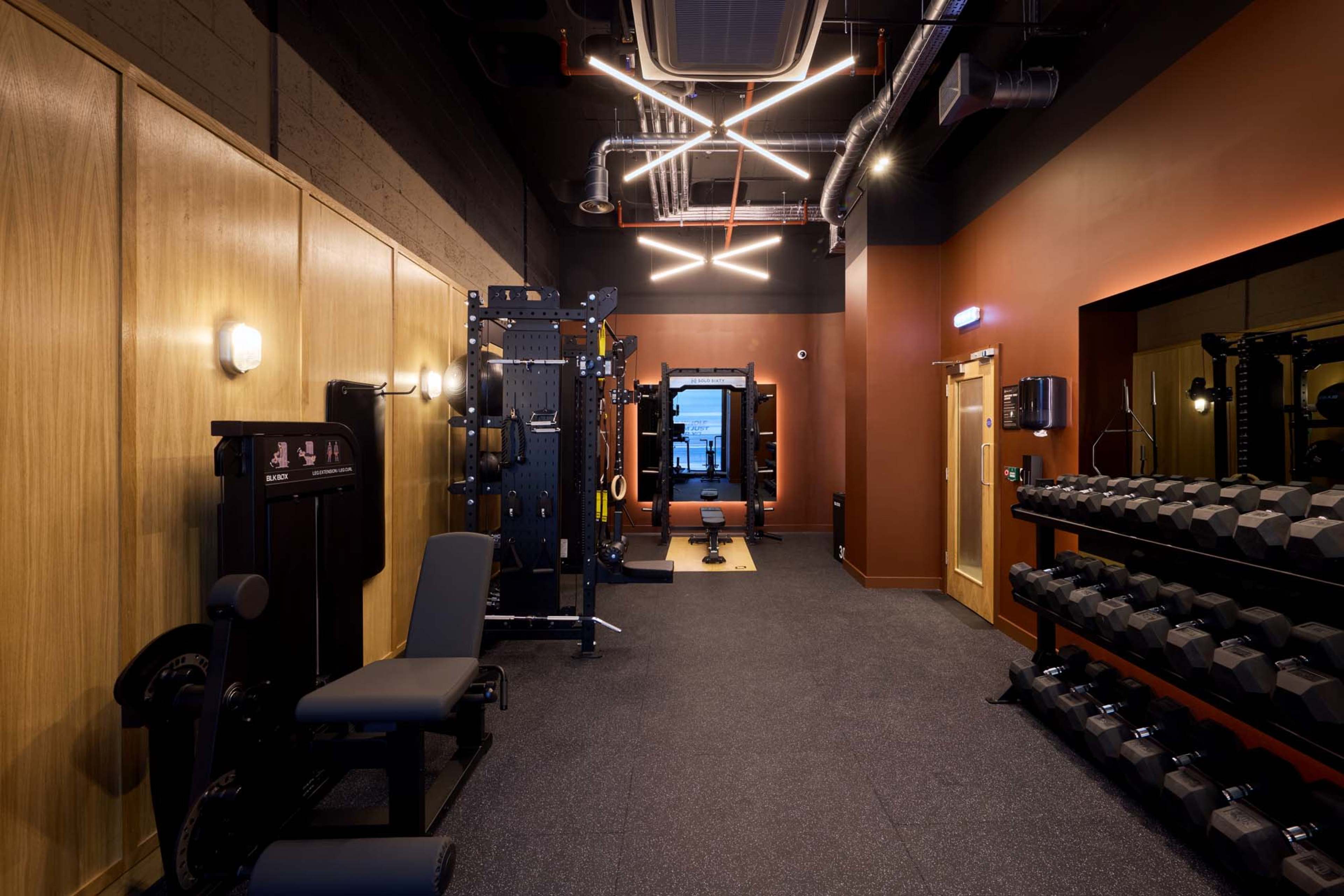 The image shows a modern gym interior featuring weightlifting equipment, dumbbells, and a mirrored wall.