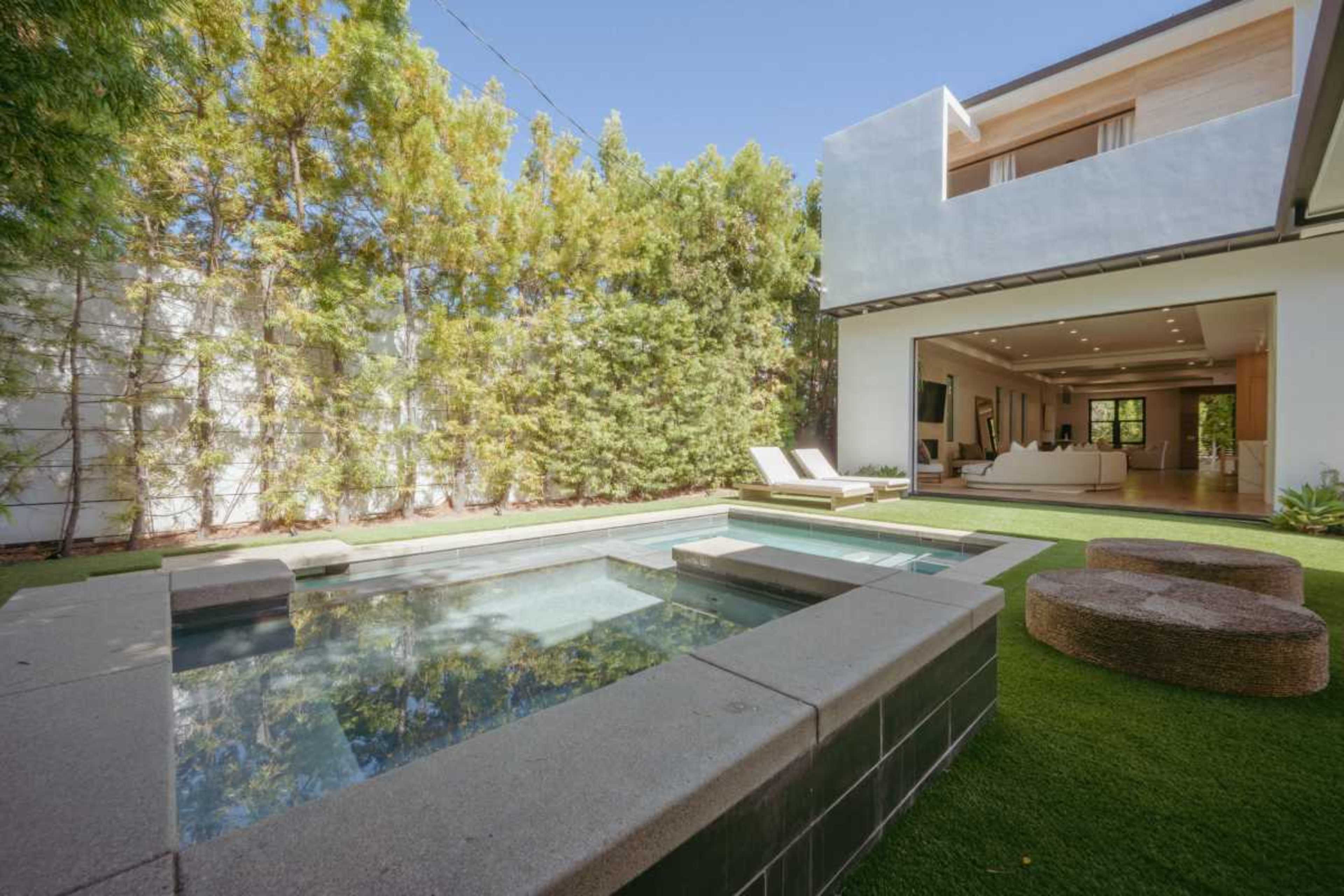 A modern backyard features a swimming pool and hot tub, surrounded by lush greenery and a contemporary home.