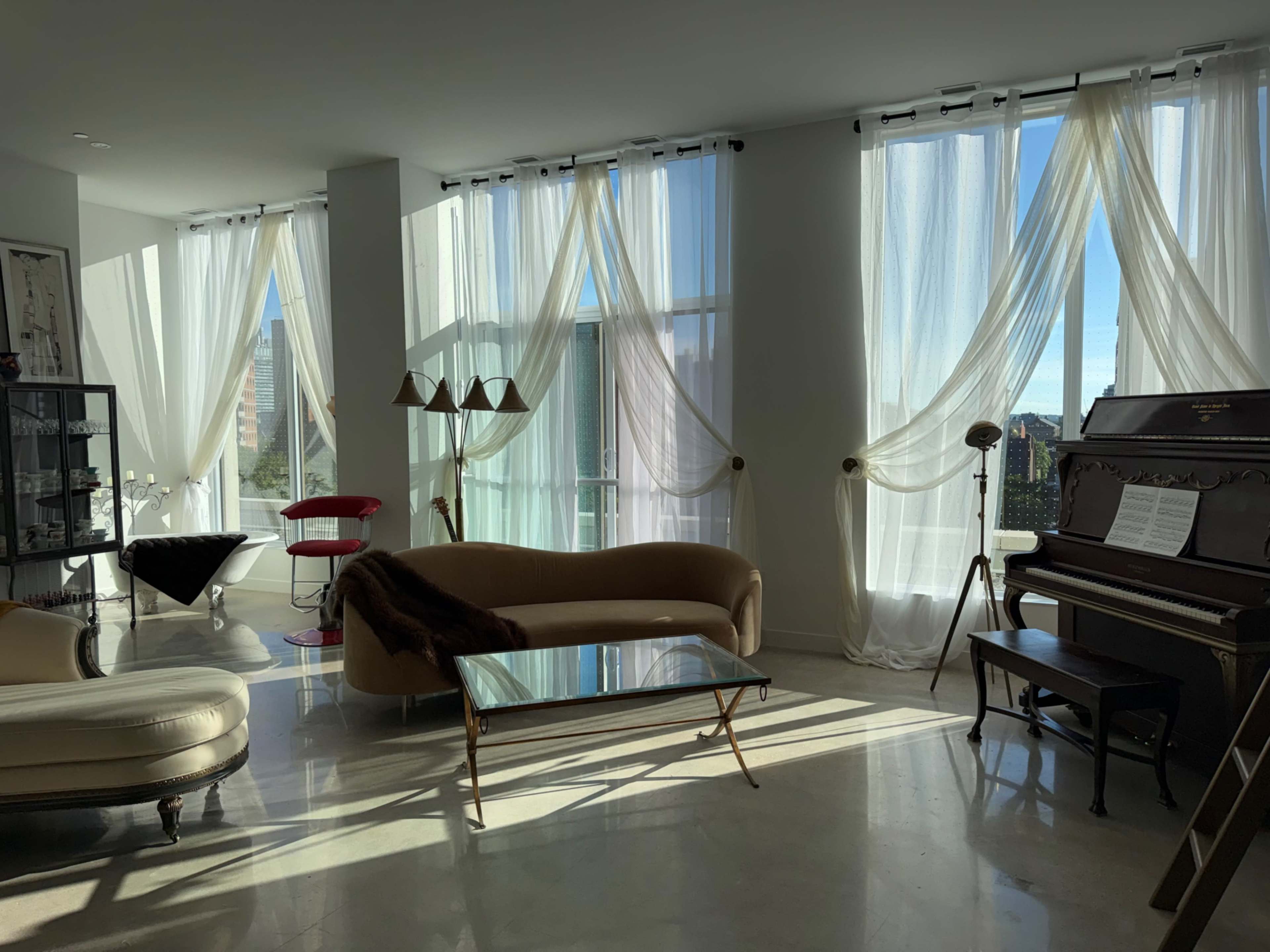 The image shows a bright room with large windows, elegant furniture, and a piano, filled with soft natural light.