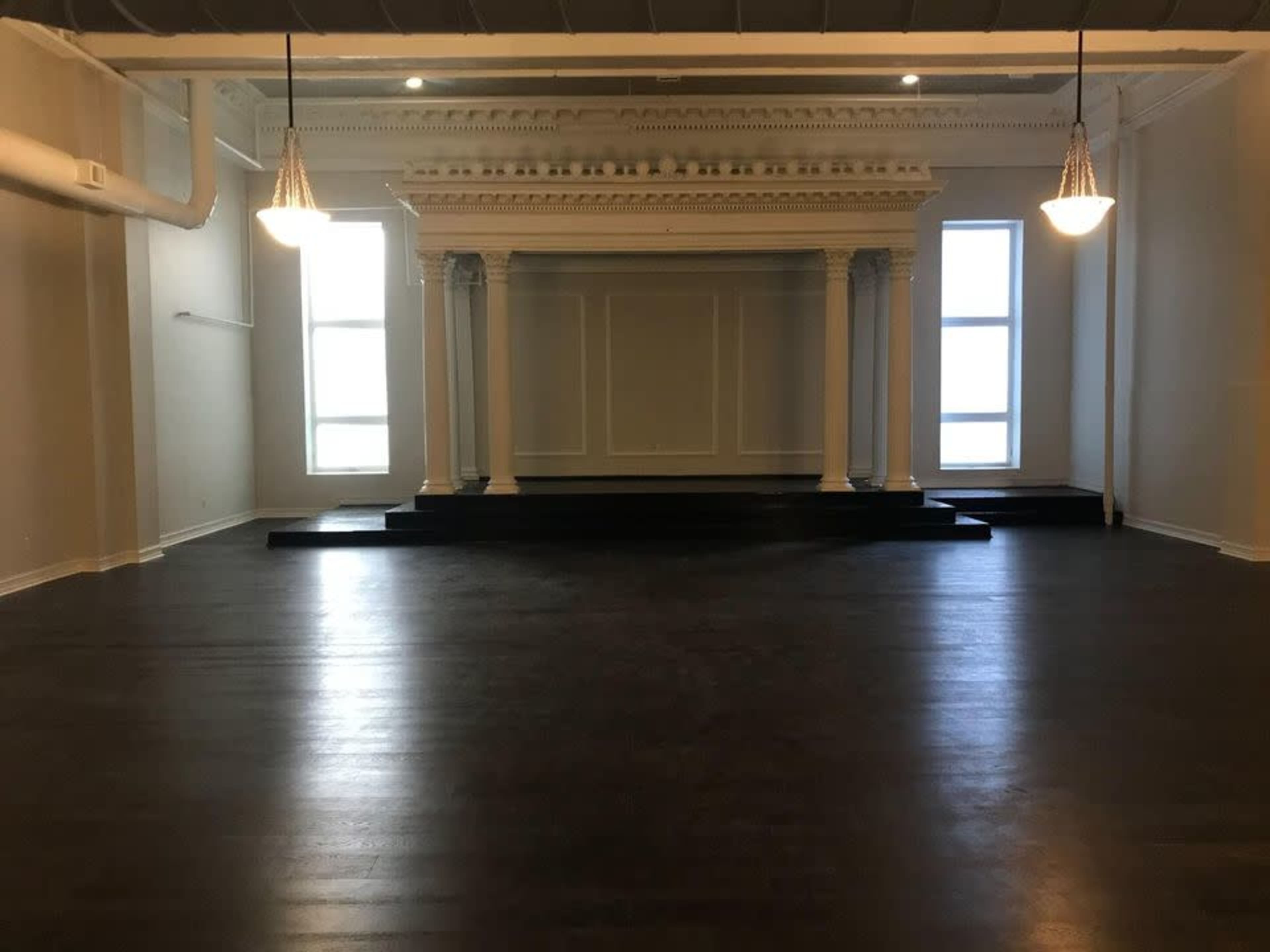 A spacious, empty room featuring a raised platform with columns and large windows at one end.