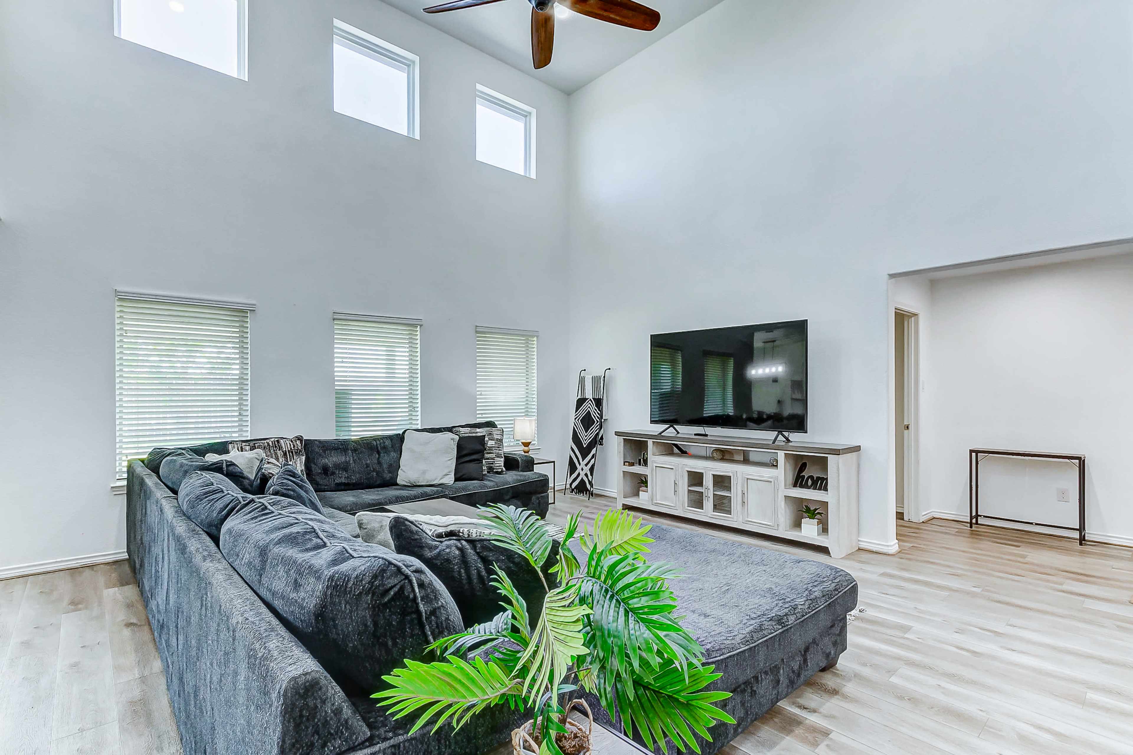 A spacious living room with large windows, a dark sectional sofa, a television on a white cabinet, and a potted plant in the corner.
