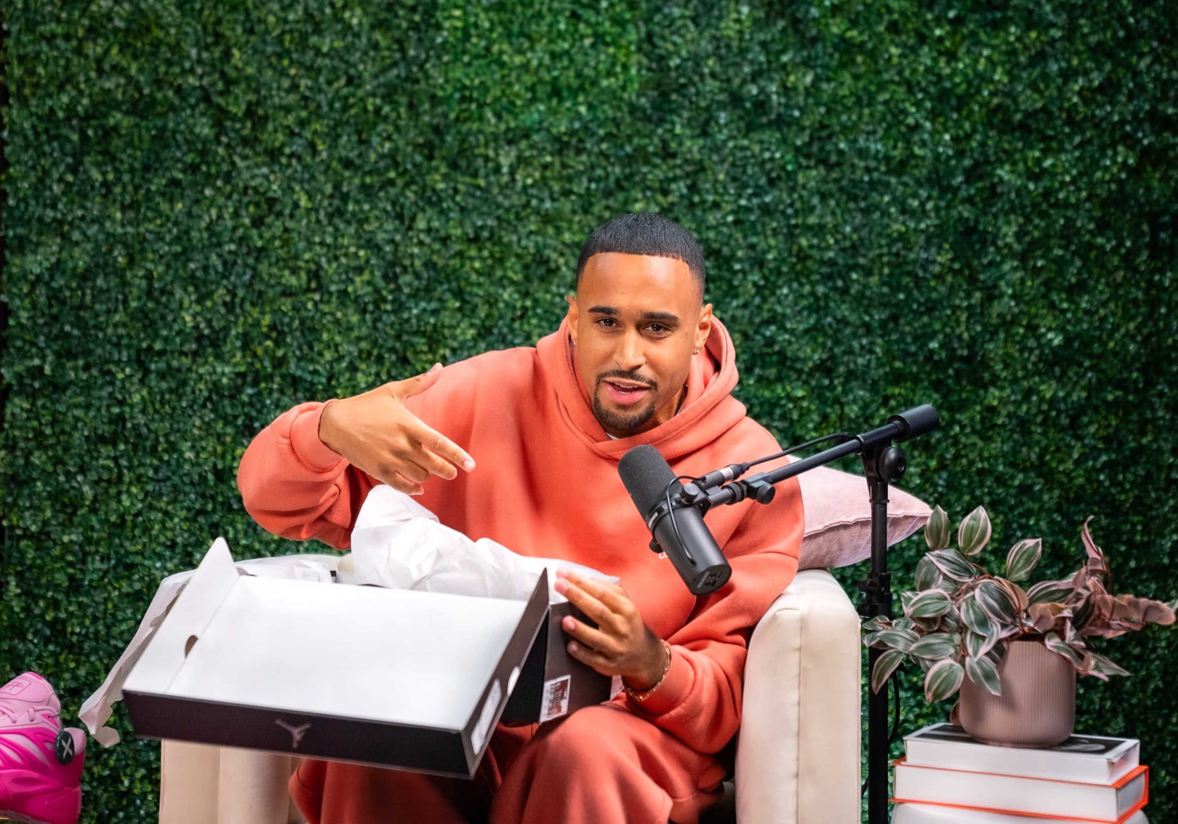A person in a pink hoodie sits on a chair, holding a shoe box while gesturing, in front of a green leafy backdrop and a microphone.