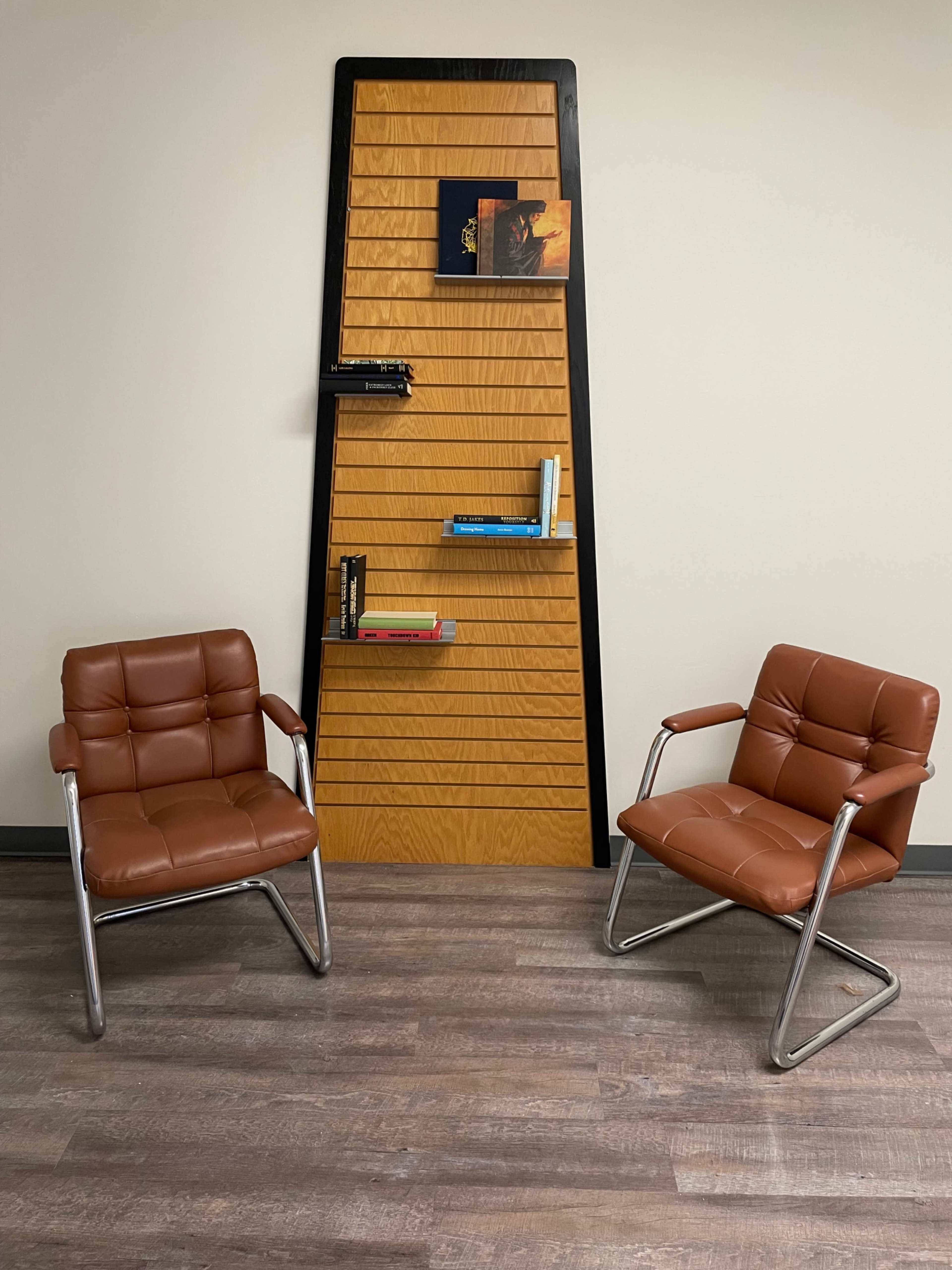 The image shows two brown chairs positioned on a wooden floor in front of a tall, decorative wooden panel with several books and a small artwork displayed.