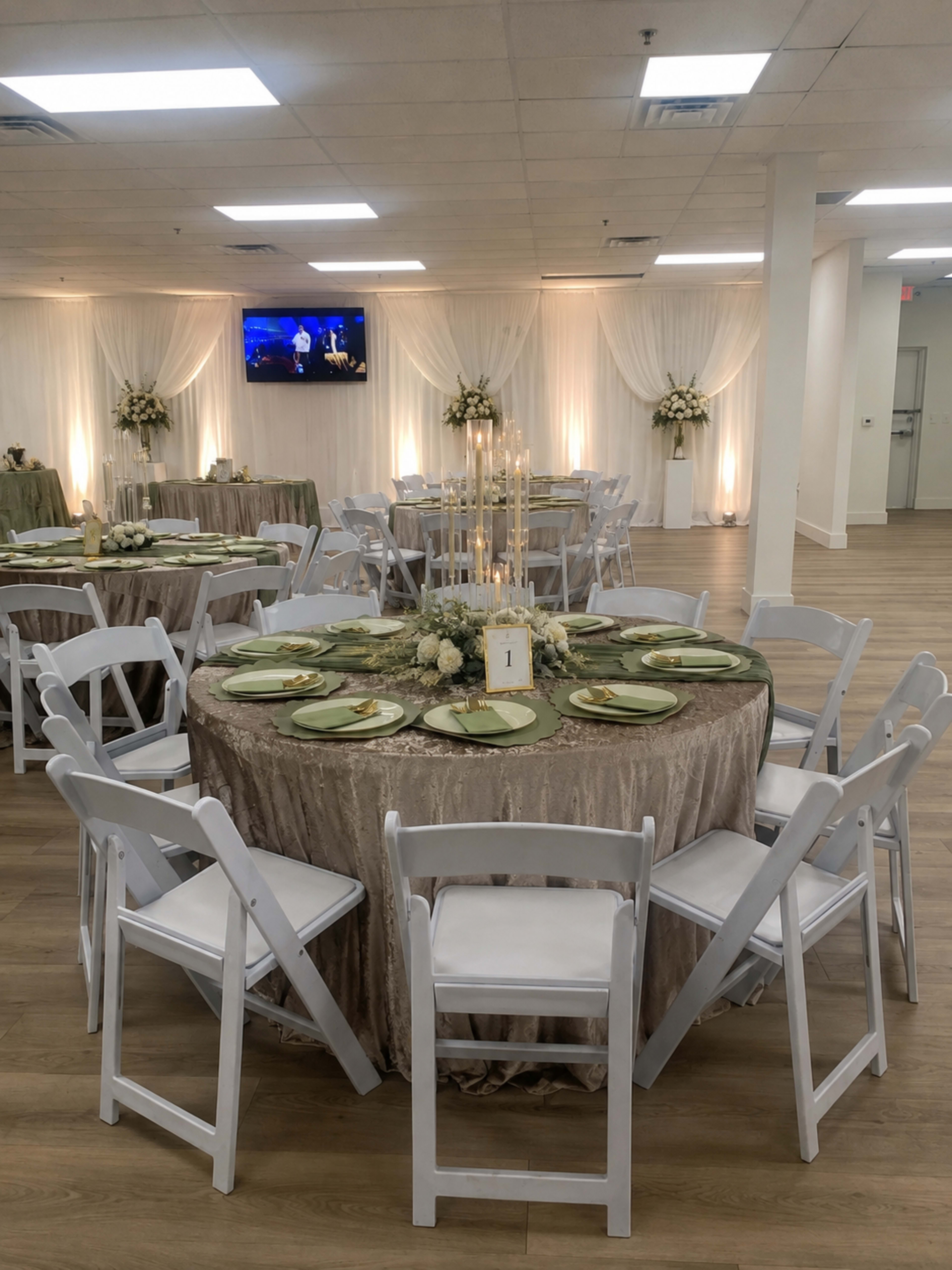 The image shows a beautifully arranged event space with tables set for a gathering, featuring elegant tablecloths, centerpieces, and candles, illuminated by soft lighting.