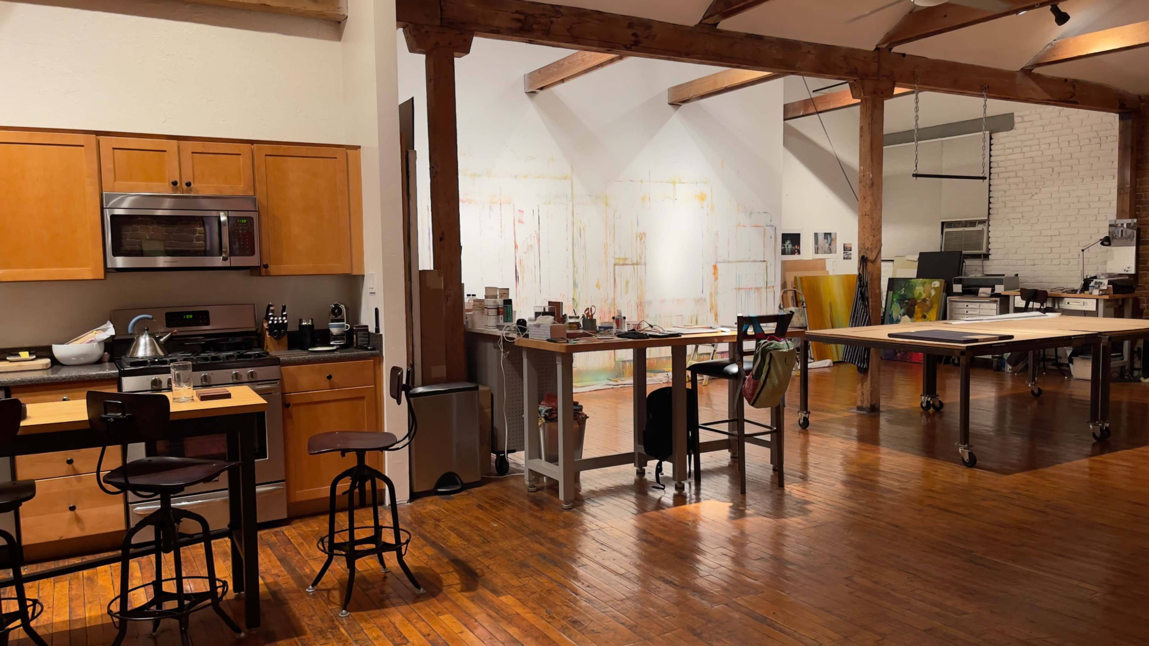 The image depicts a spacious artist's studio with wooden beams, a kitchen area with cabinets, and various workstations arranged in an open layout.