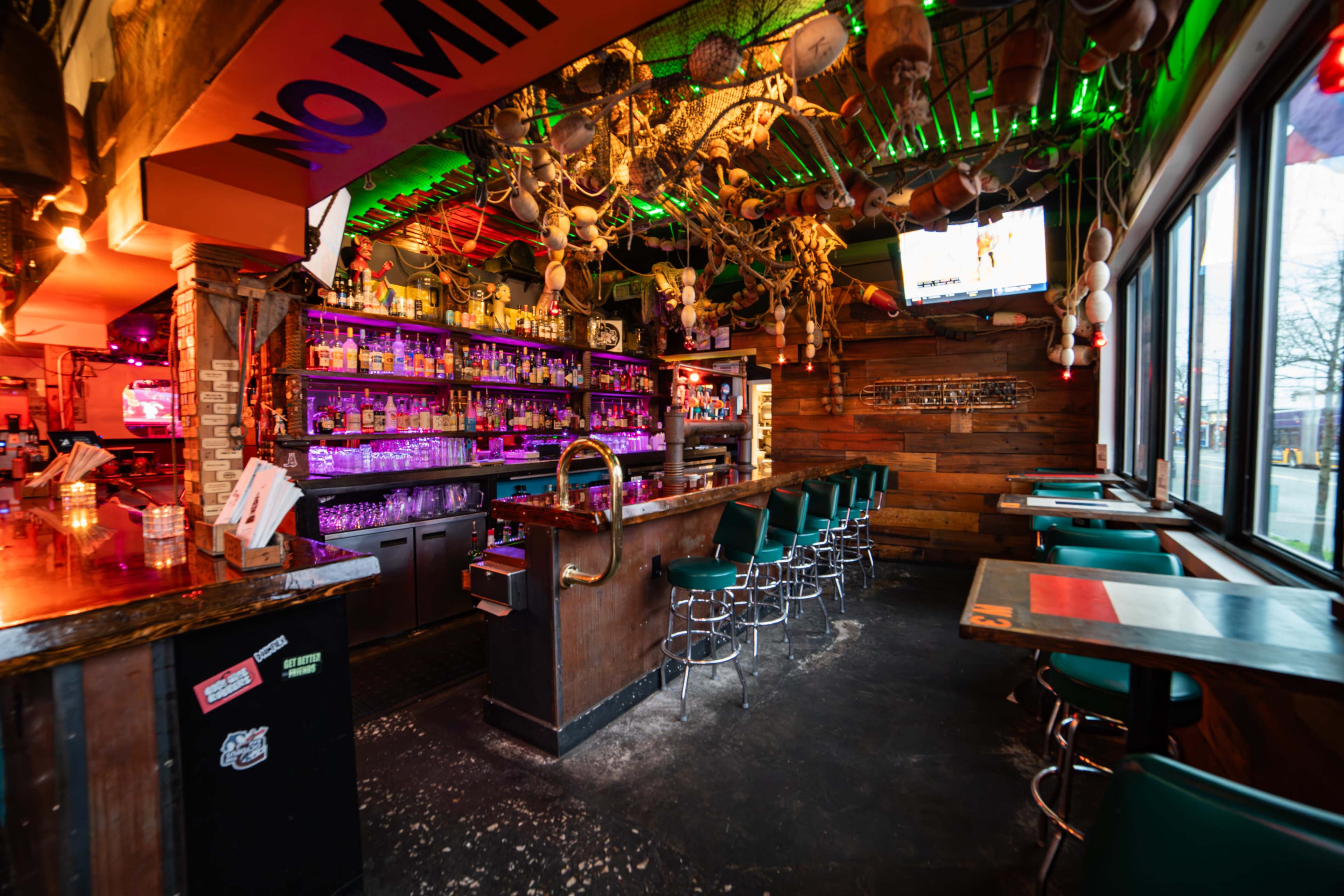 The interior of a bar features a long wooden counter, high stools, shelves filled with colorful bottles, and lively decorations hanging from the ceiling.