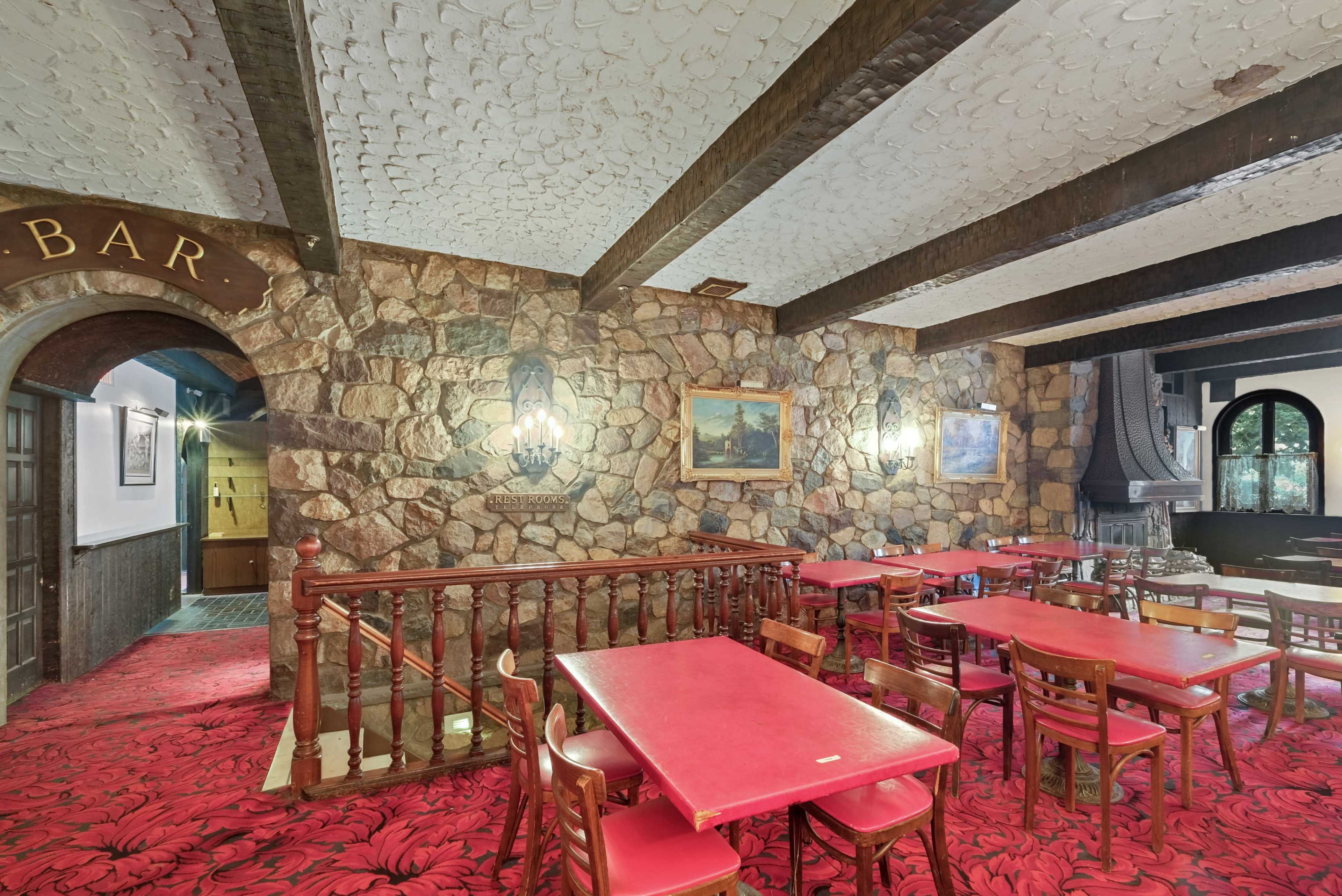A rustic bar area featuring stone walls, wooden beams, red-topped tables, and a patterned carpet.