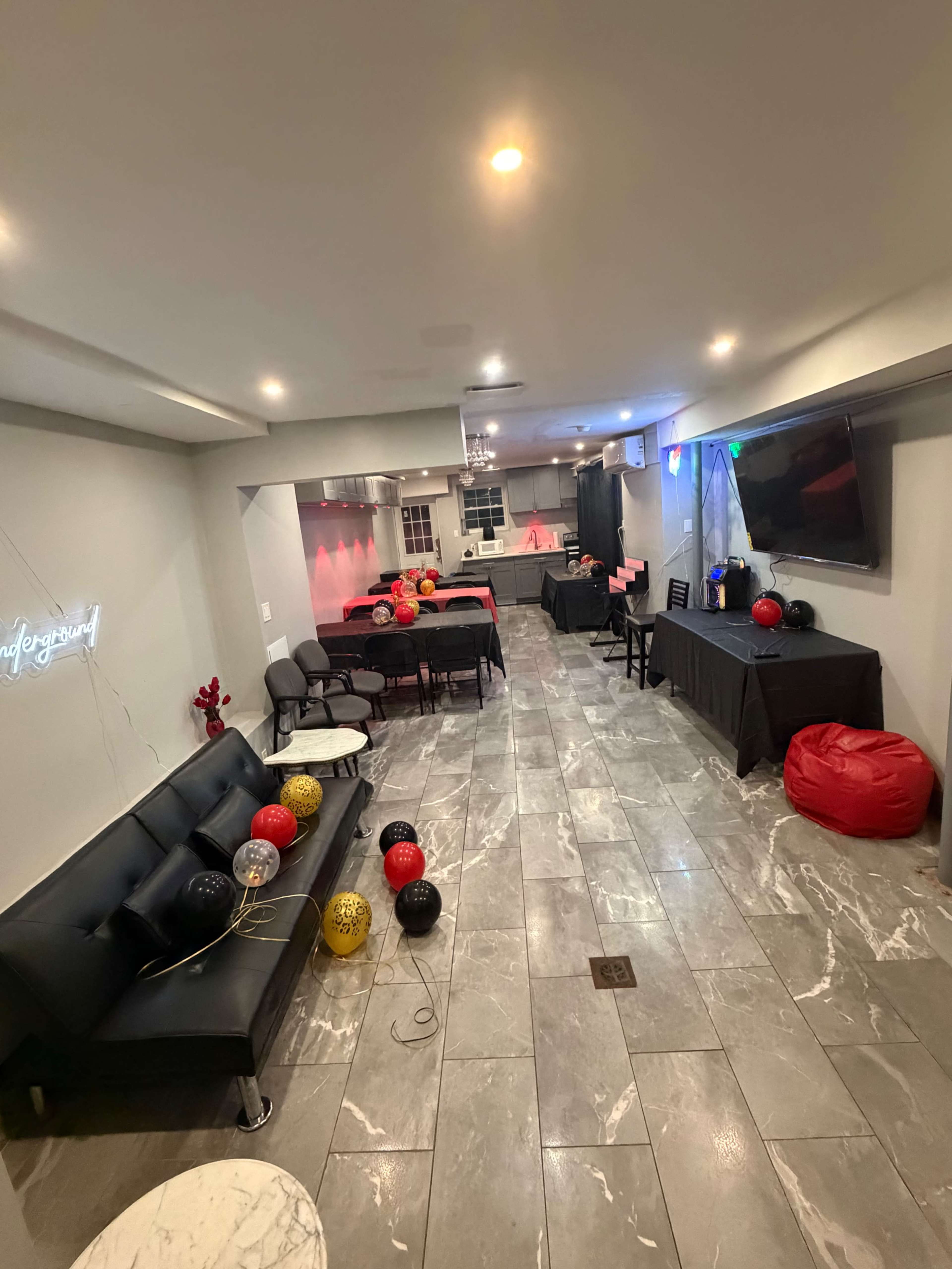 The image shows a decorated indoor event space with black and red balloons, tables covered with black cloths, and seating areas featuring a black couch and bean bags.