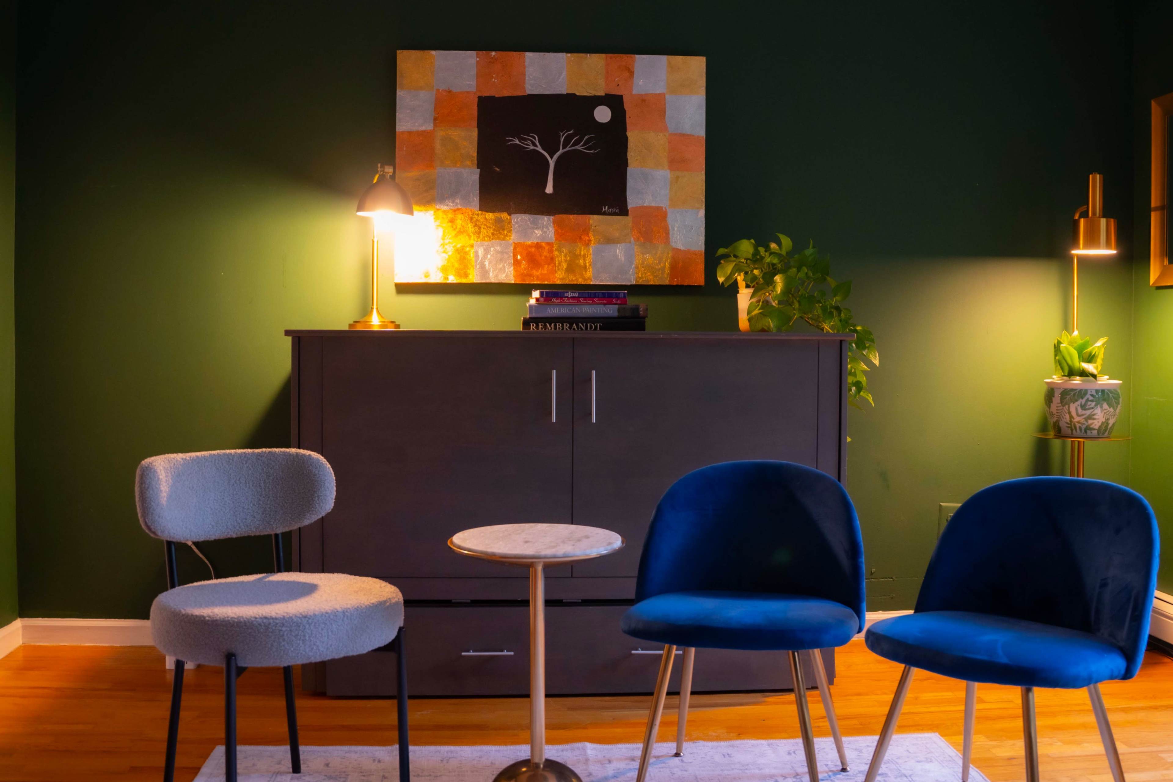 The image shows a cozy living room with a dark green wall, a wooden cabinet, three chairs in blue and grey, a small round table, and a piece of artwork above the cabinet.