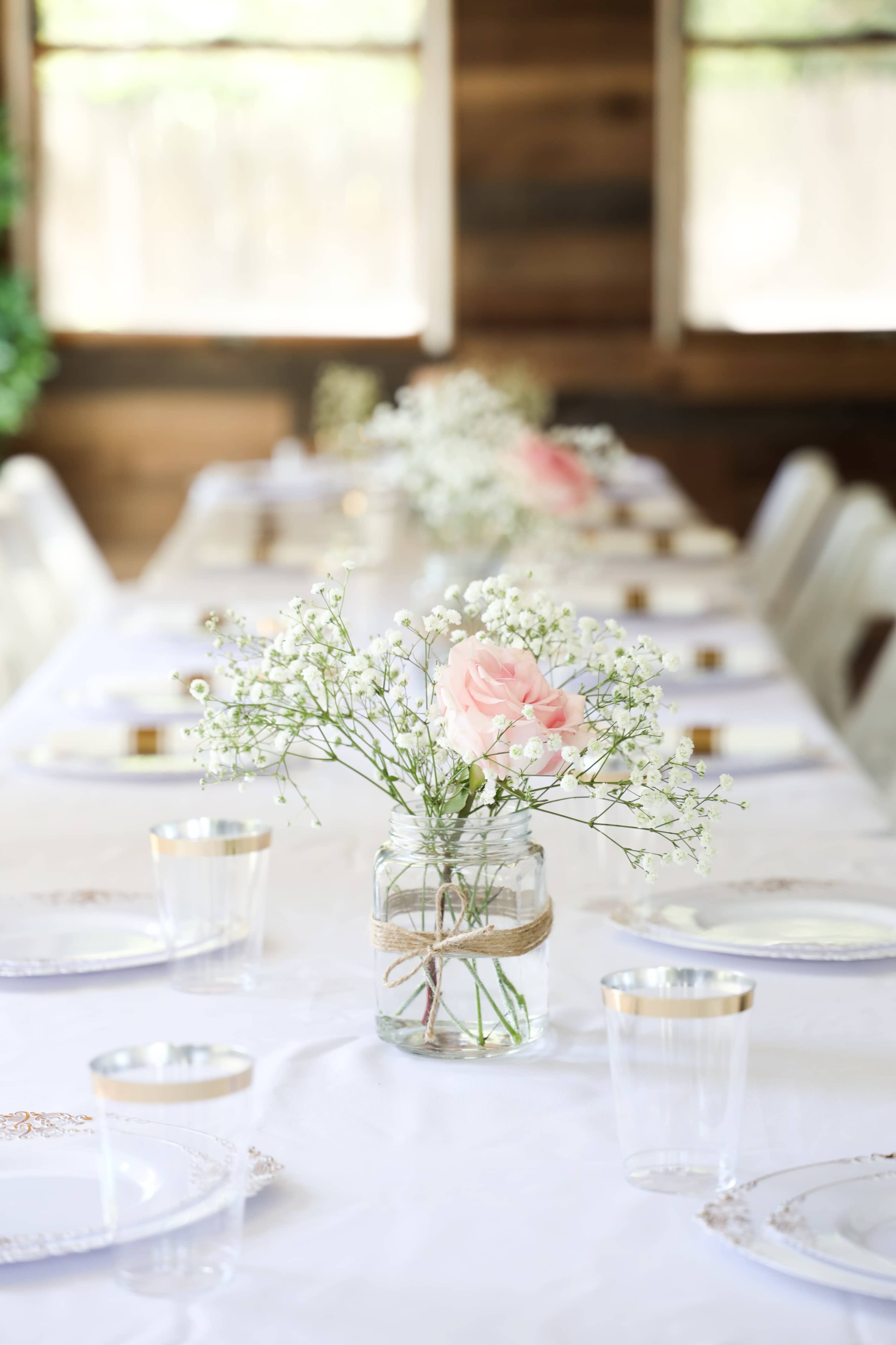 A long table is set for a gathering, adorned with a white tablecloth, clear glasses, and jars of flowers as centerpieces.