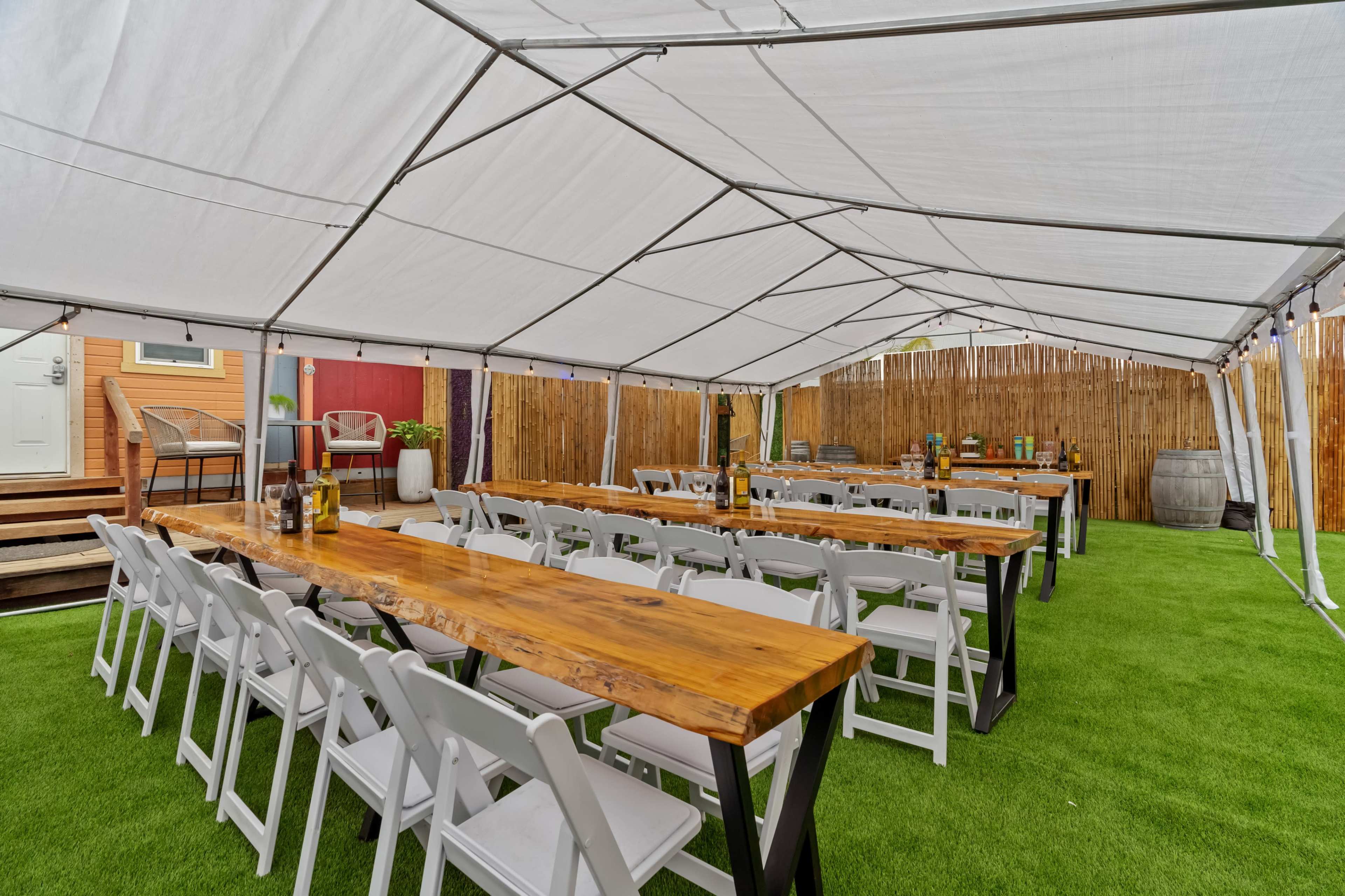 A large outdoor tent is set up with long wooden tables and white chairs arranged neatly on green artificial grass.