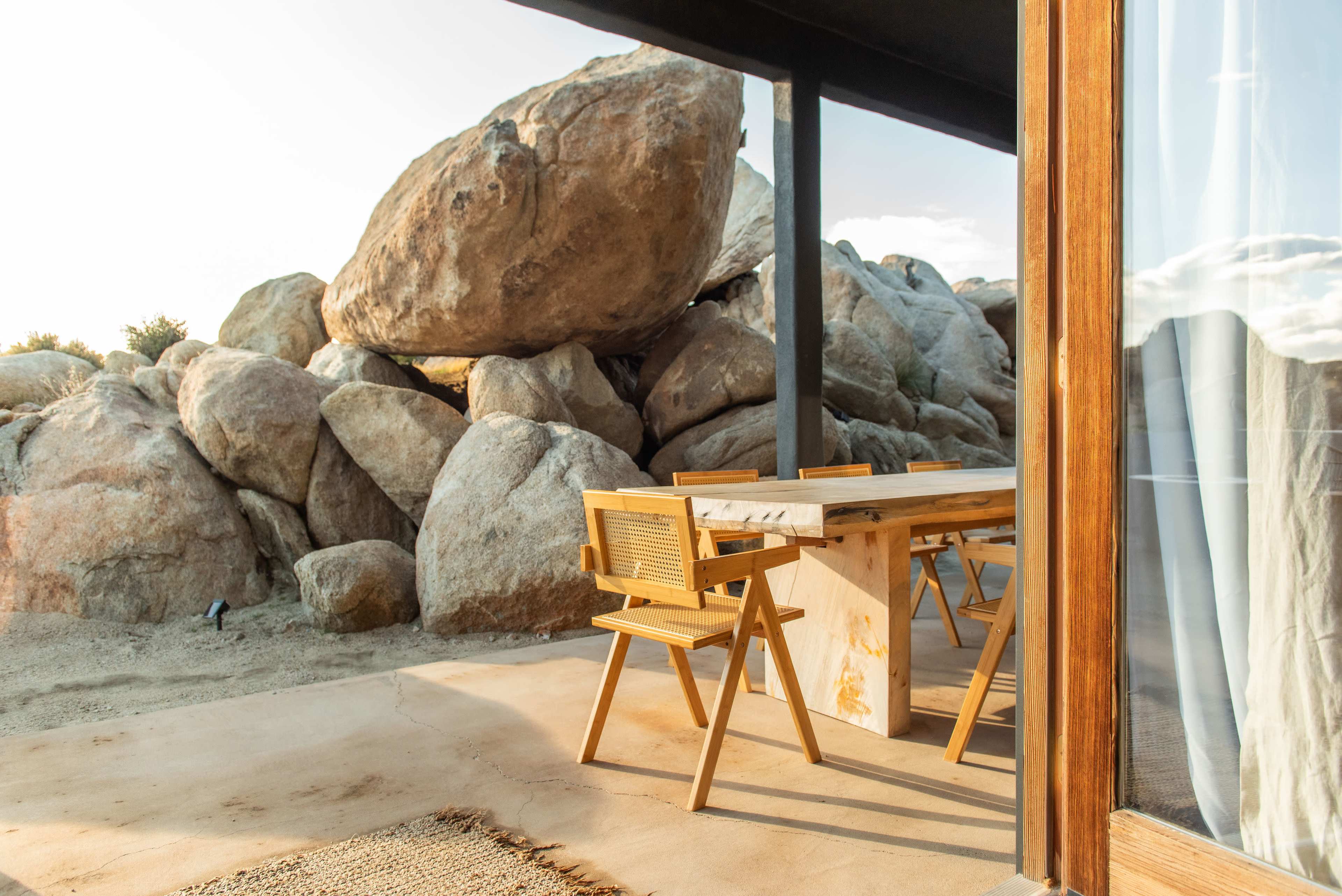 A wooden dining table with chairs is positioned outside a structure, рядом with large boulders in a natural landscape.