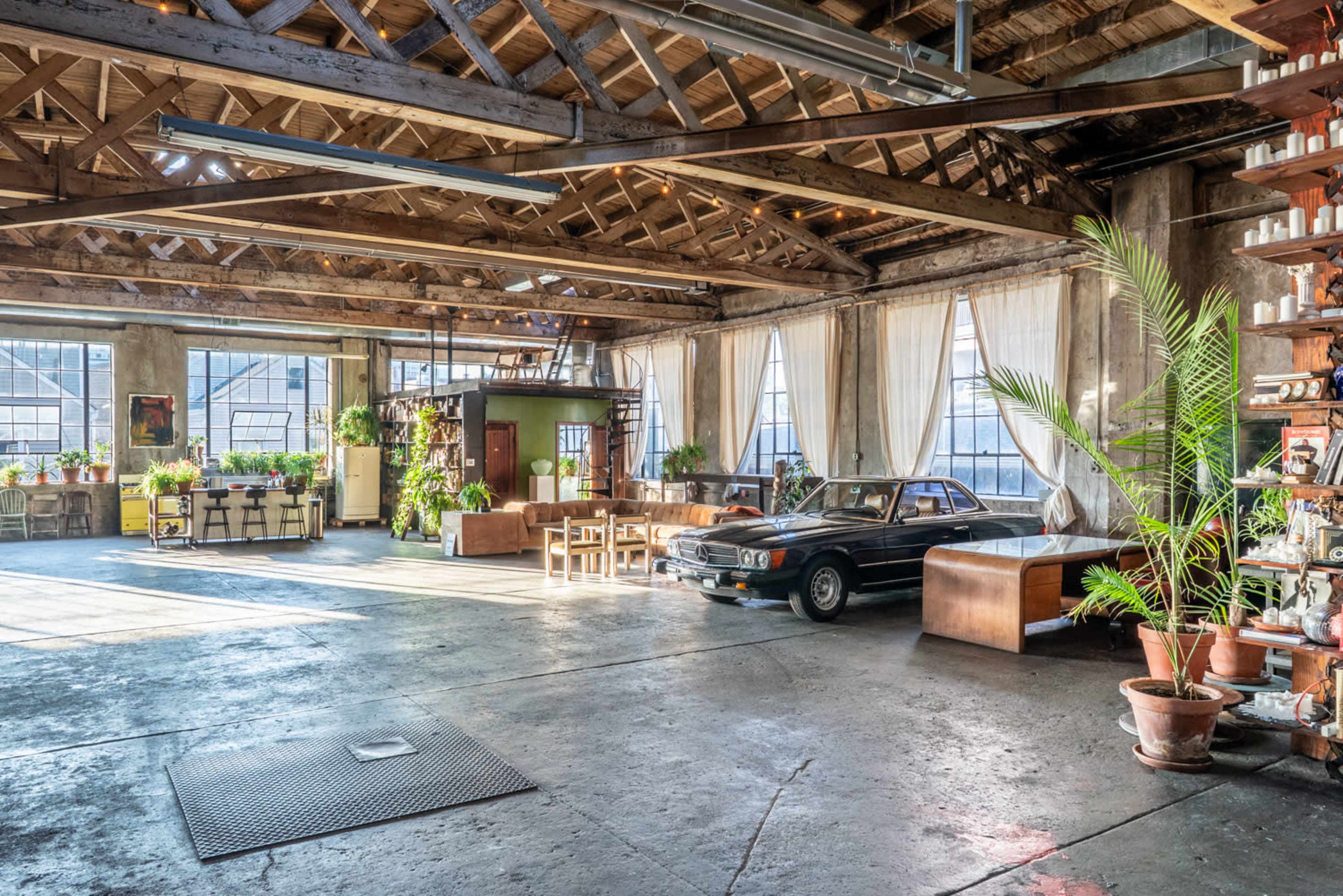 The image shows a spacious industrial loft with exposed wooden beams, large windows, a classic car parked in the center, and various plants arranged around the area.