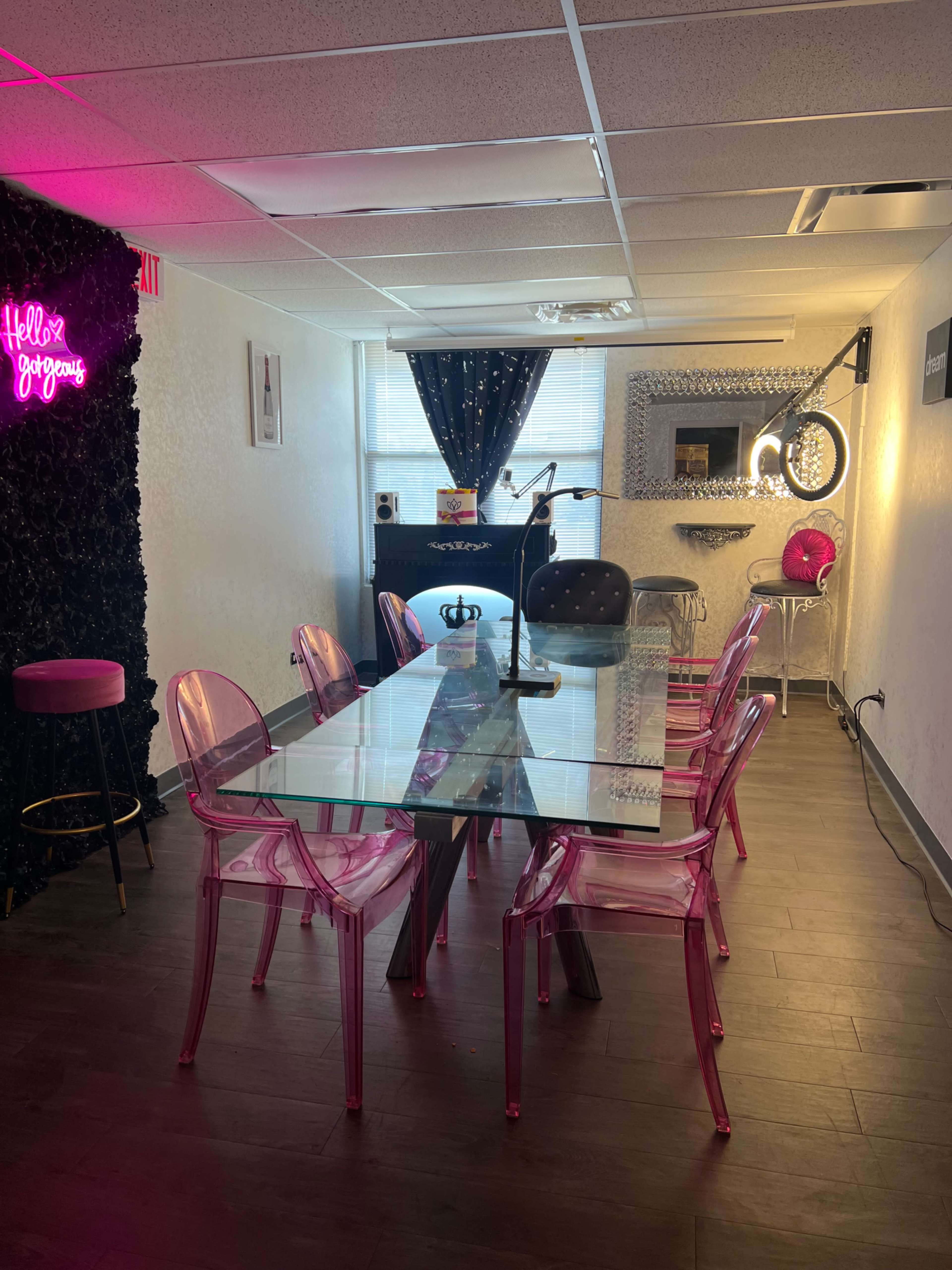 The image shows a modern meeting room with a glass table surrounded by transparent pink chairs, decorated with a black textured wall and ambient lighting.
