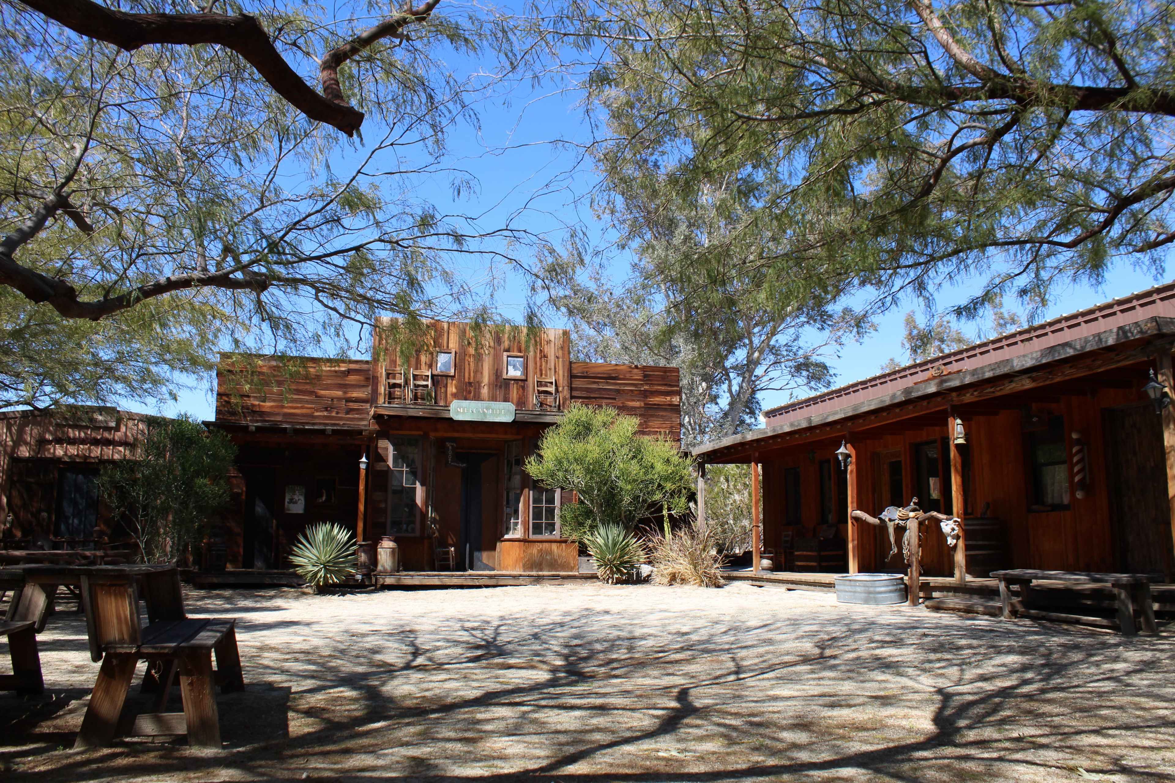 An old western-style building surrounded by sparse trees and a gravel courtyard, with wooden structures on either side.