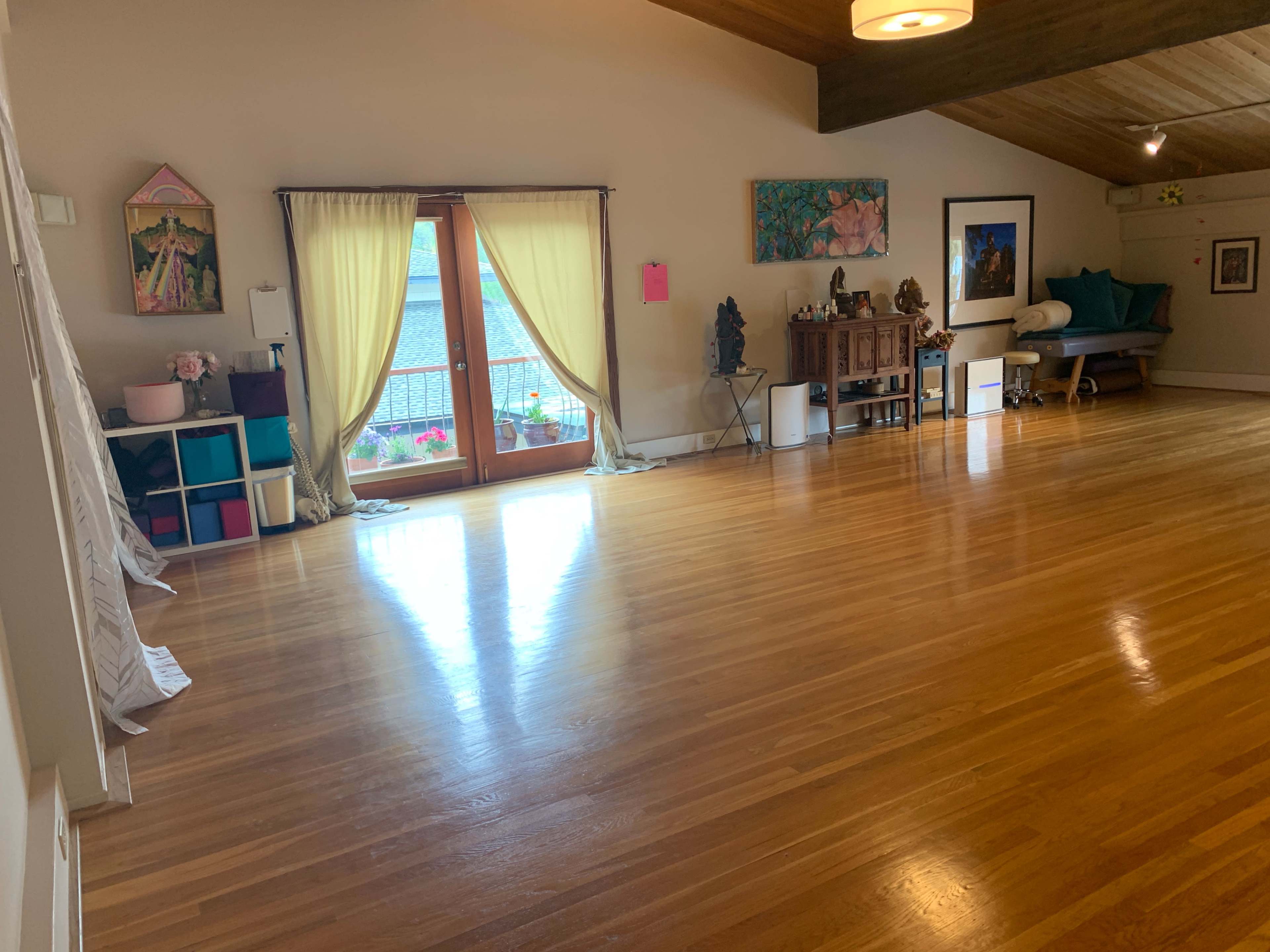 A spacious, well-lit room with wooden flooring, large windows, and various decorations, including artwork and furniture along the walls.