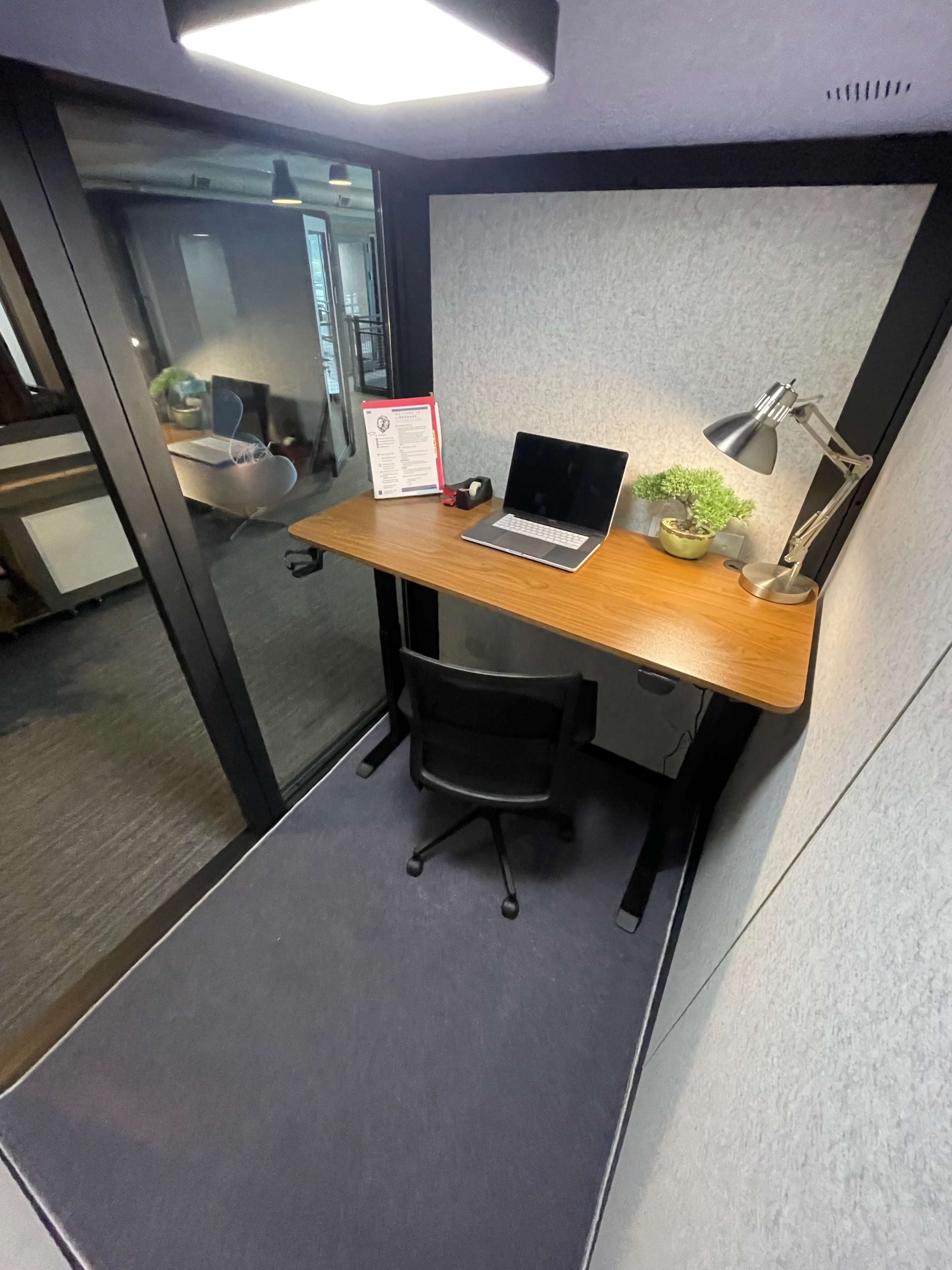 The image shows a small office nook featuring a desk with a laptop, a lamp, and a small plant, enclosed by soundproof walls and a glass door.