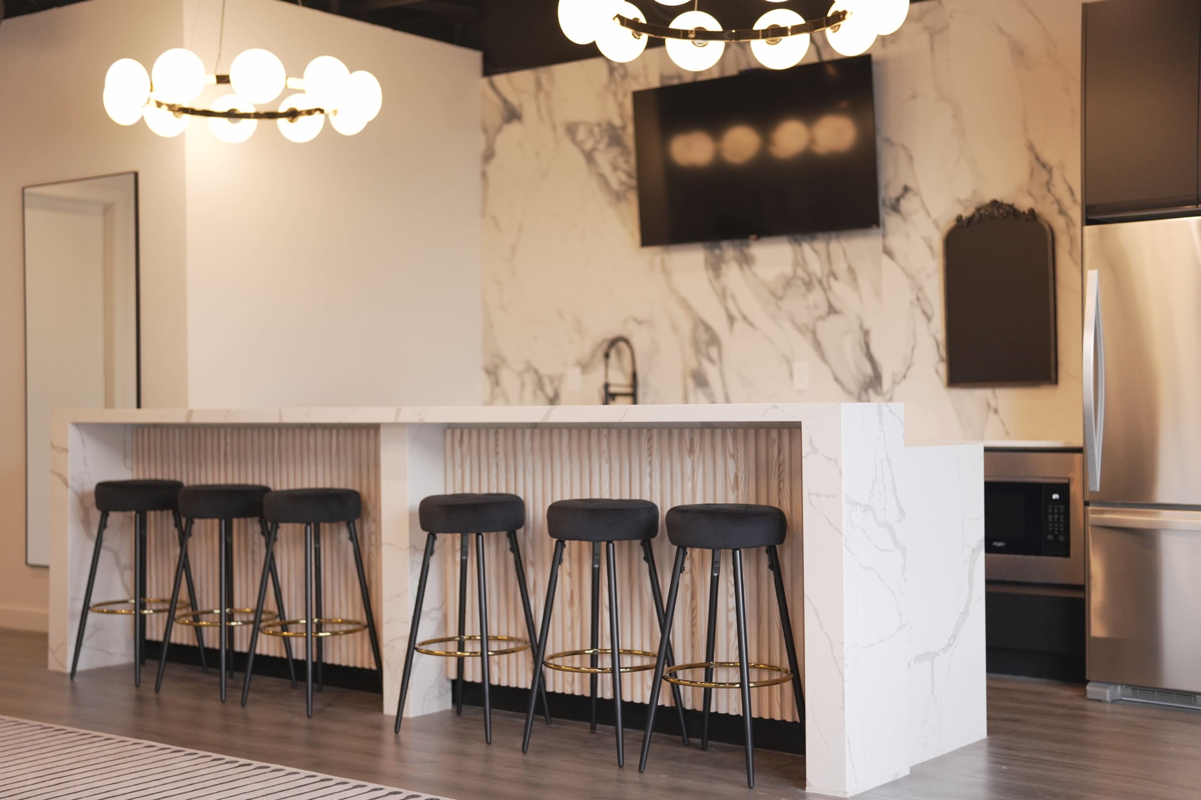A modern kitchen features a marble bar with six black stools and a large flat-screen television on the wall.