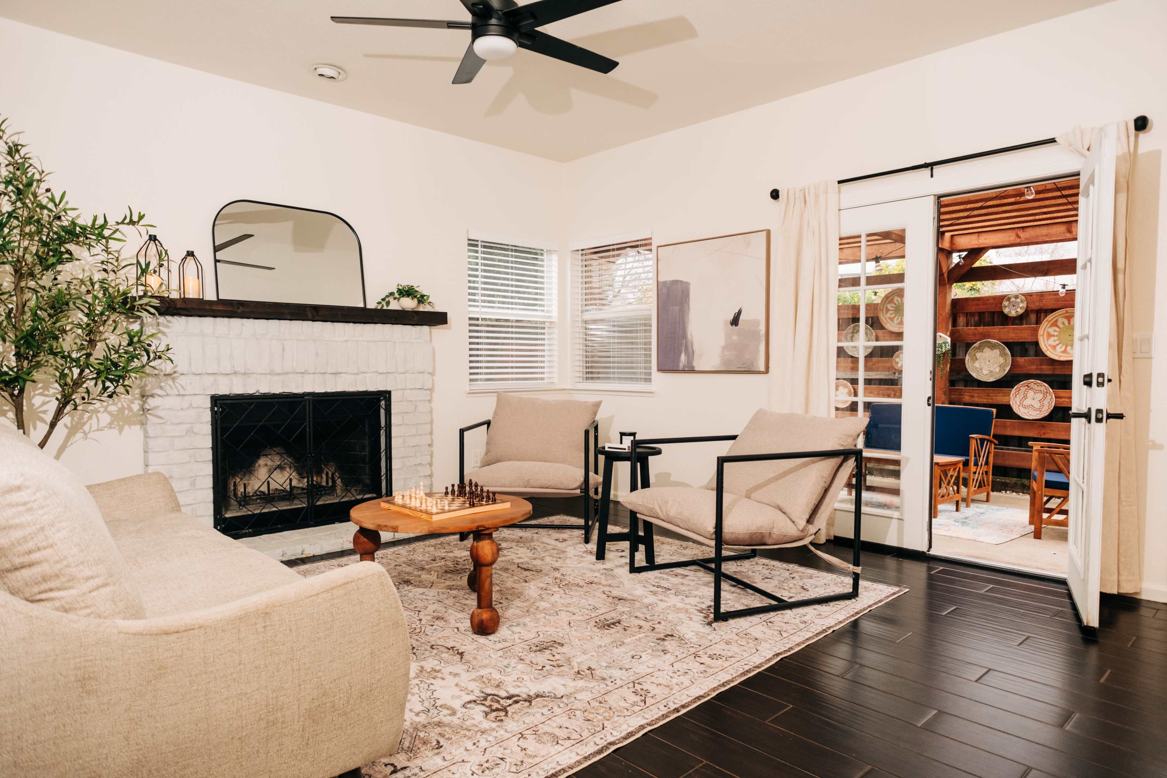 A modern living room features a fireplace, a chess set on a wooden table, two armchairs, and sliding glass doors leading to an outdoor area.