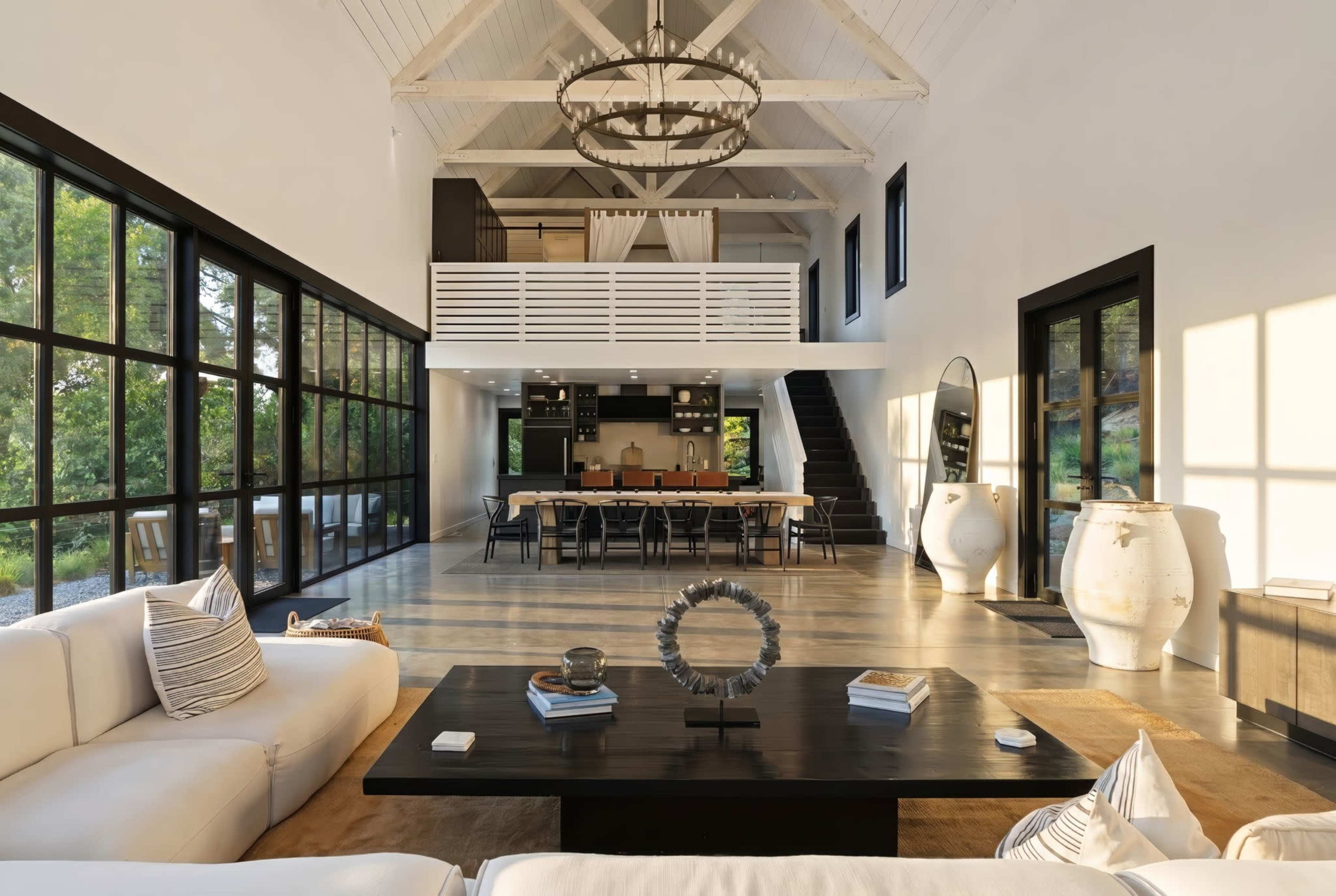 The image shows a modern open-concept living area with high ceilings, large windows, and a dining table set for multiple people.