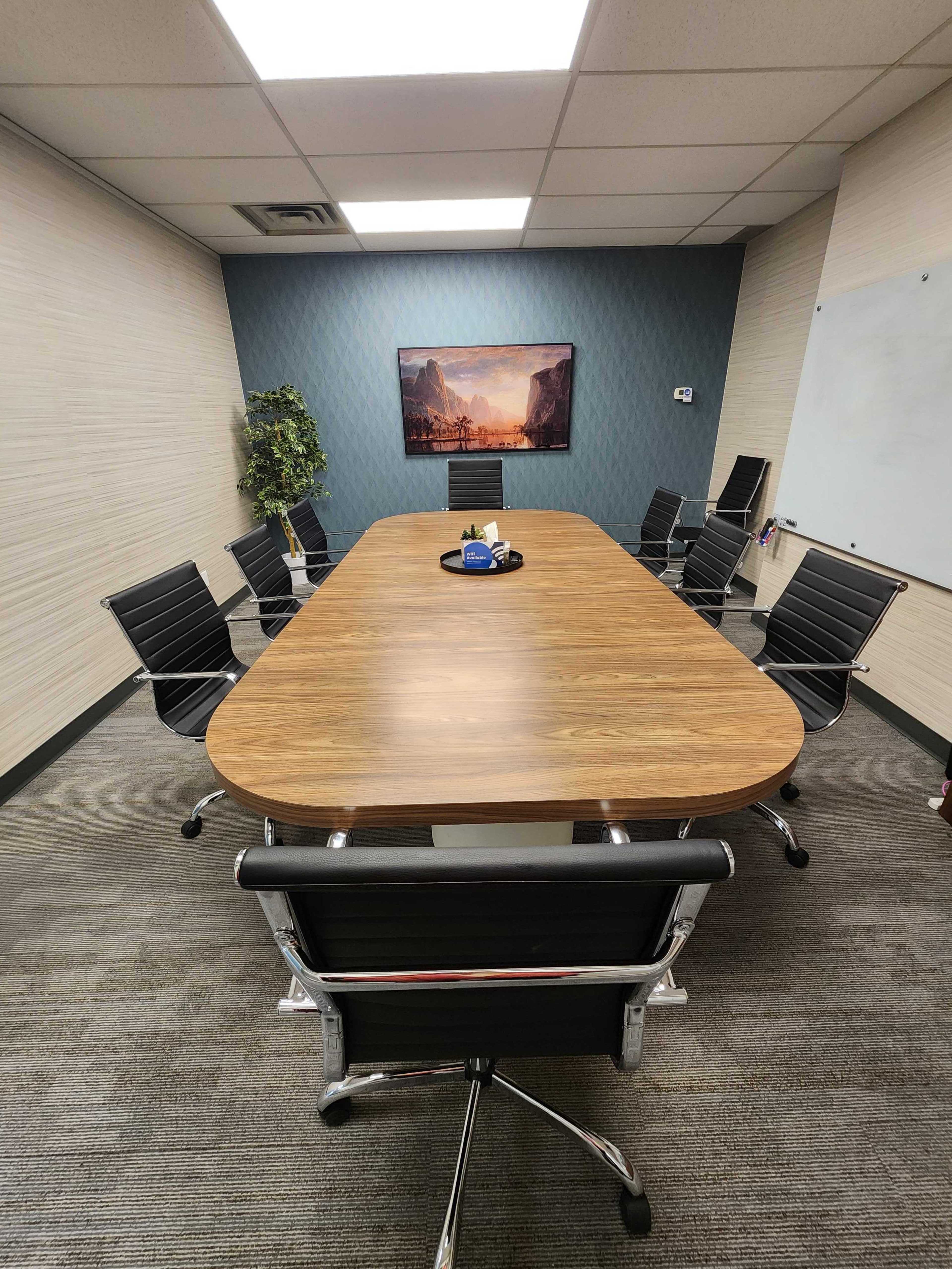A rectangular wooden conference table surrounded by black office chairs is set in a meeting room with a plant and a wall-mounted picture in the background.