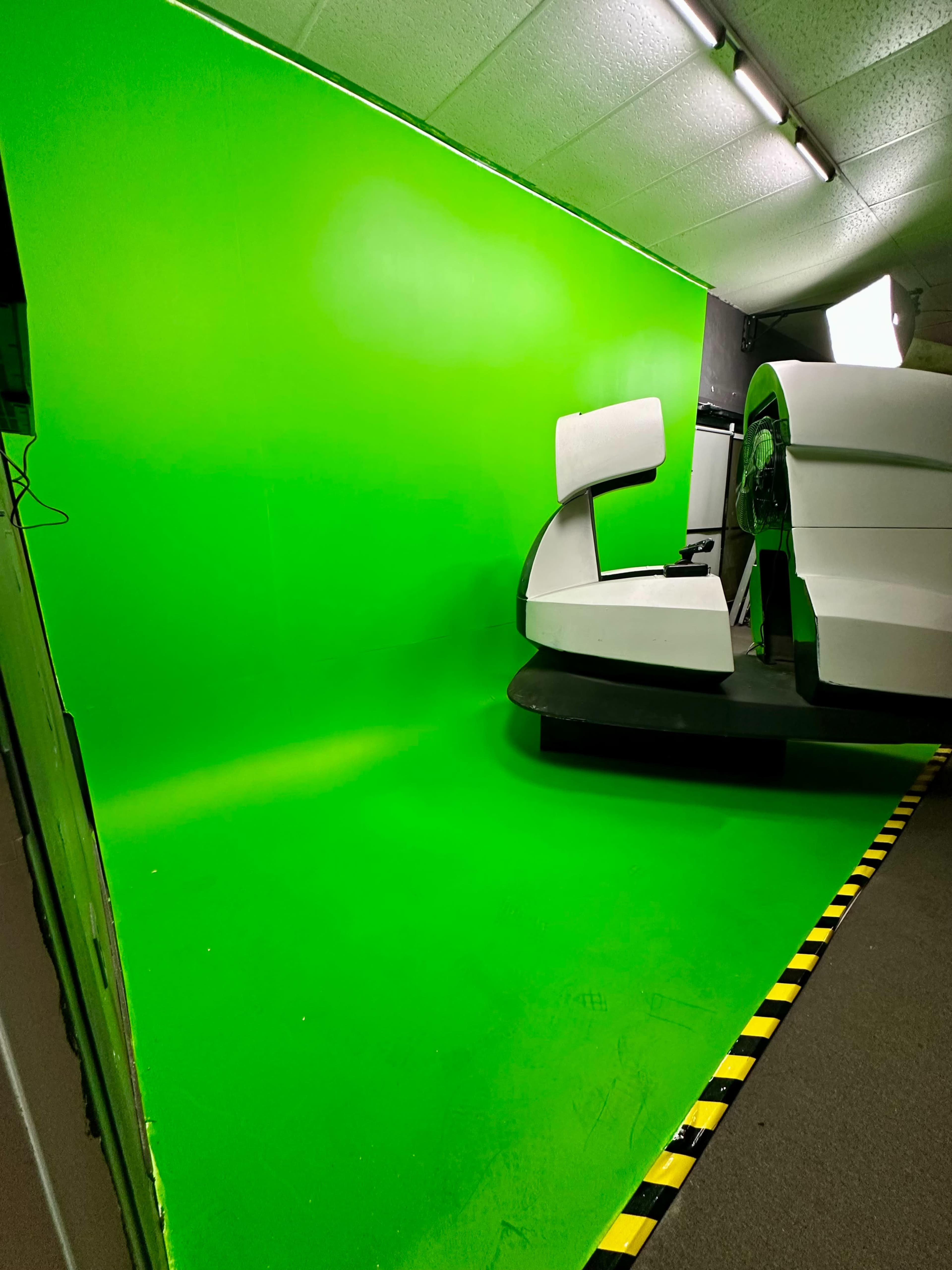 The image shows a green screen setup with a white, modern chair positioned in front of it, along with a spotlight in the corner.