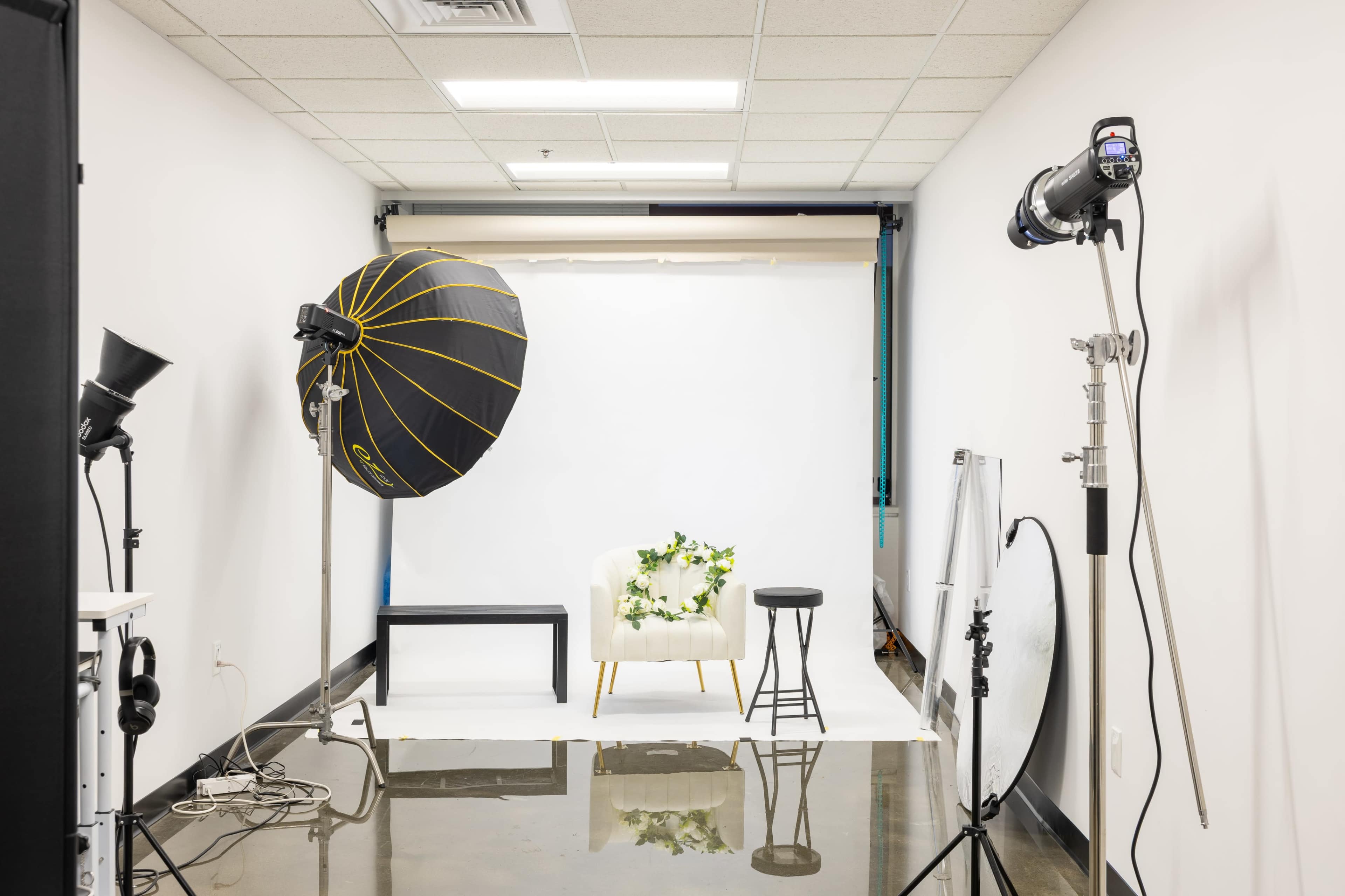 The image shows a photography studio with a white backdrop, a chair adorned with flowers, and various lighting equipment arranged around the space.