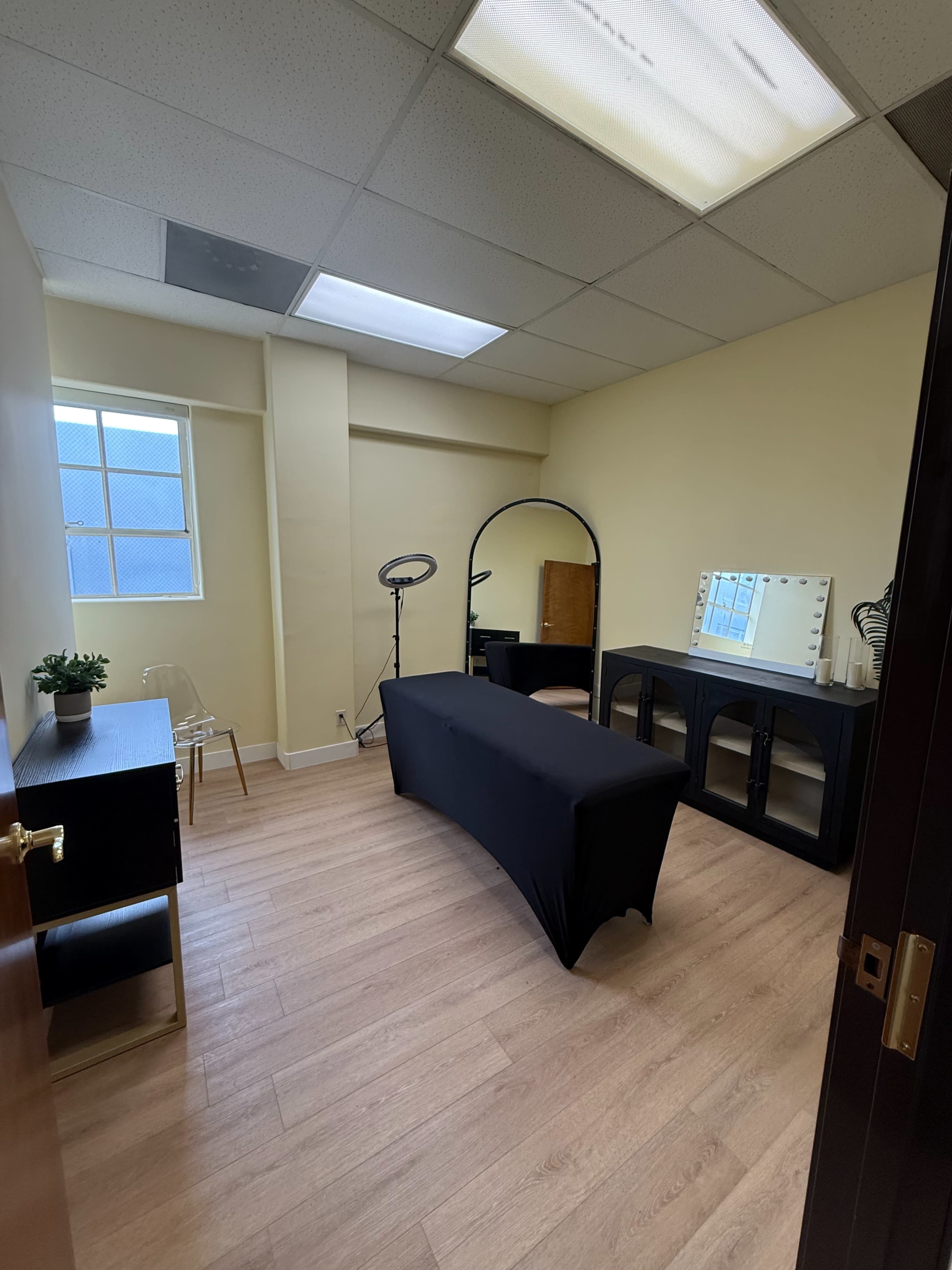 The image shows a small, well-lit room furnished with a black table, a mirror, and a chair, with light wood flooring and beige walls.