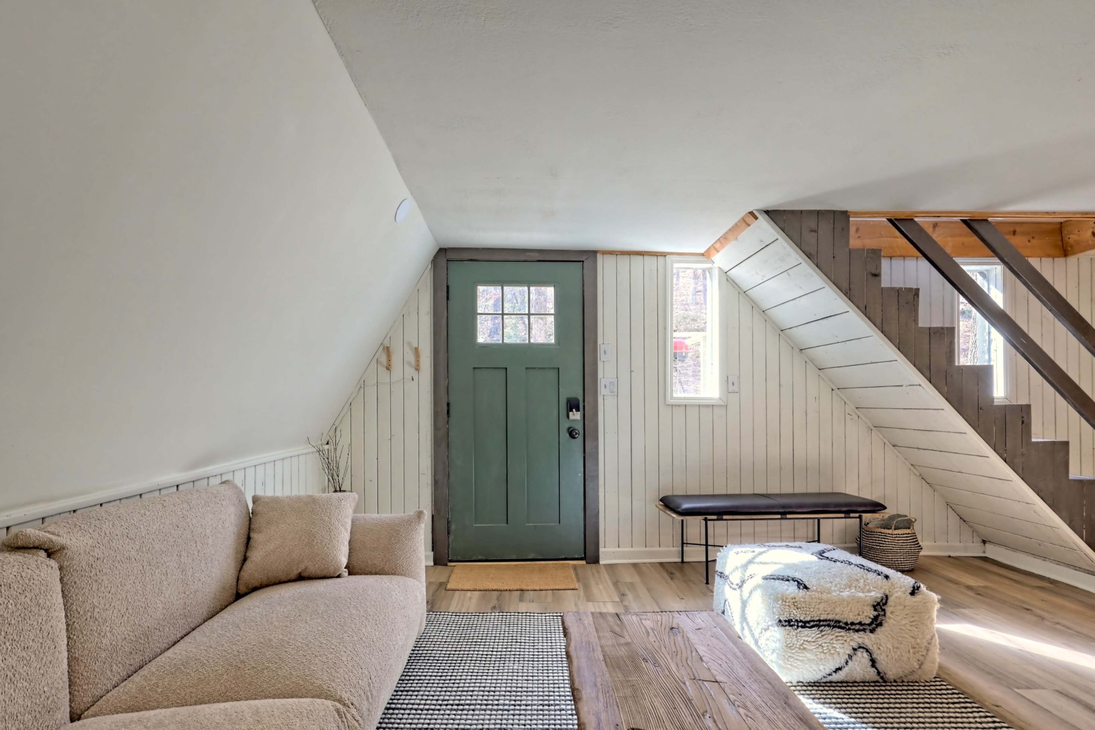 The interior of an attic space features a green door, a light-colored sofa, a wooden coffee table, and a staircase leading to the upper level.