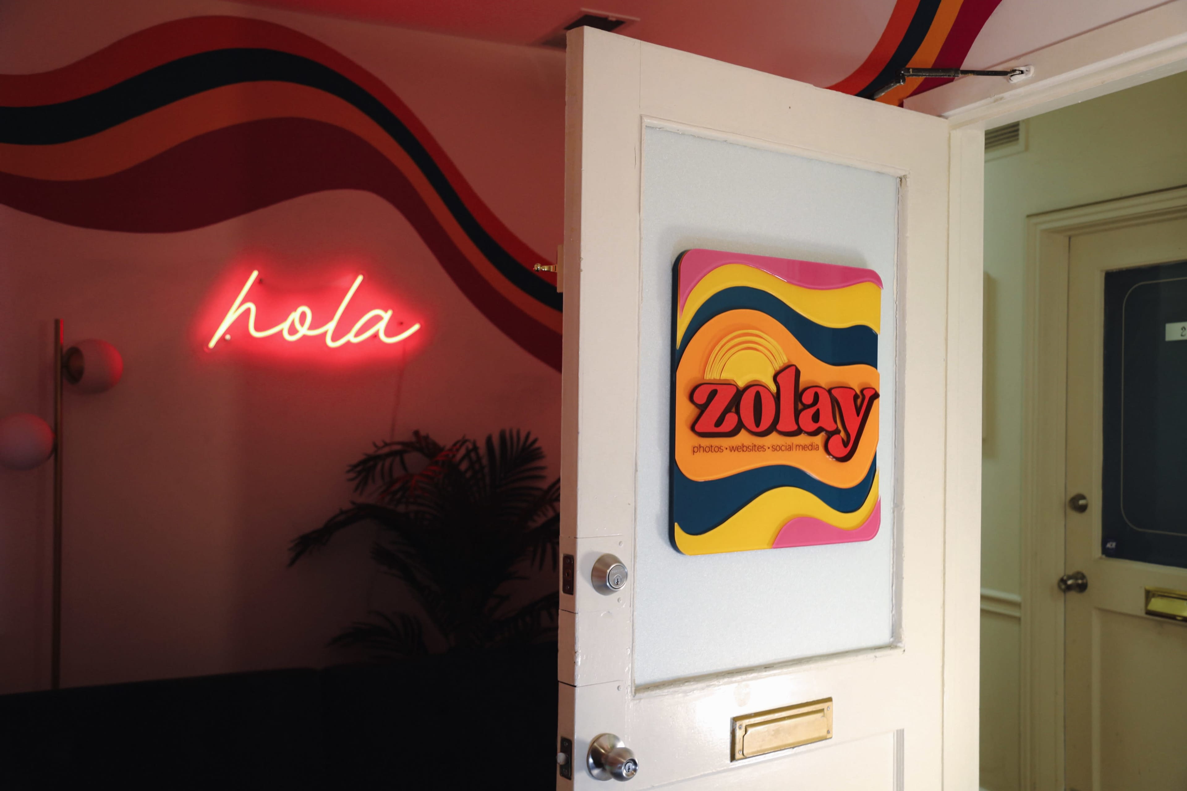 A door partially open reveals a colorful wall with the words "hola" in neon and a sign that reads "zolay" on the adjacent wall.
