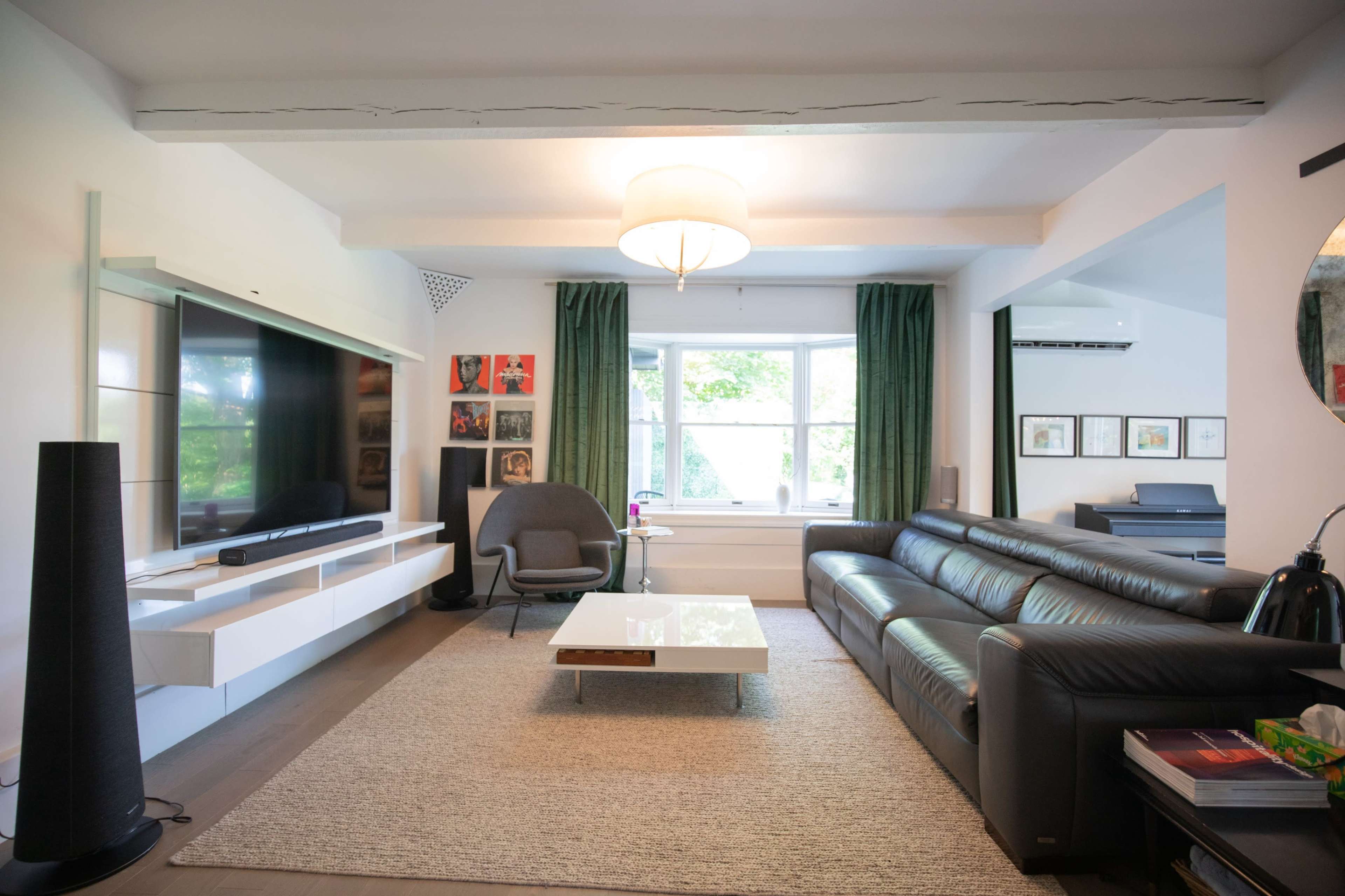 The image shows a modern living room featuring a large window, a black leather sofa, a coffee table, and a mounted television.