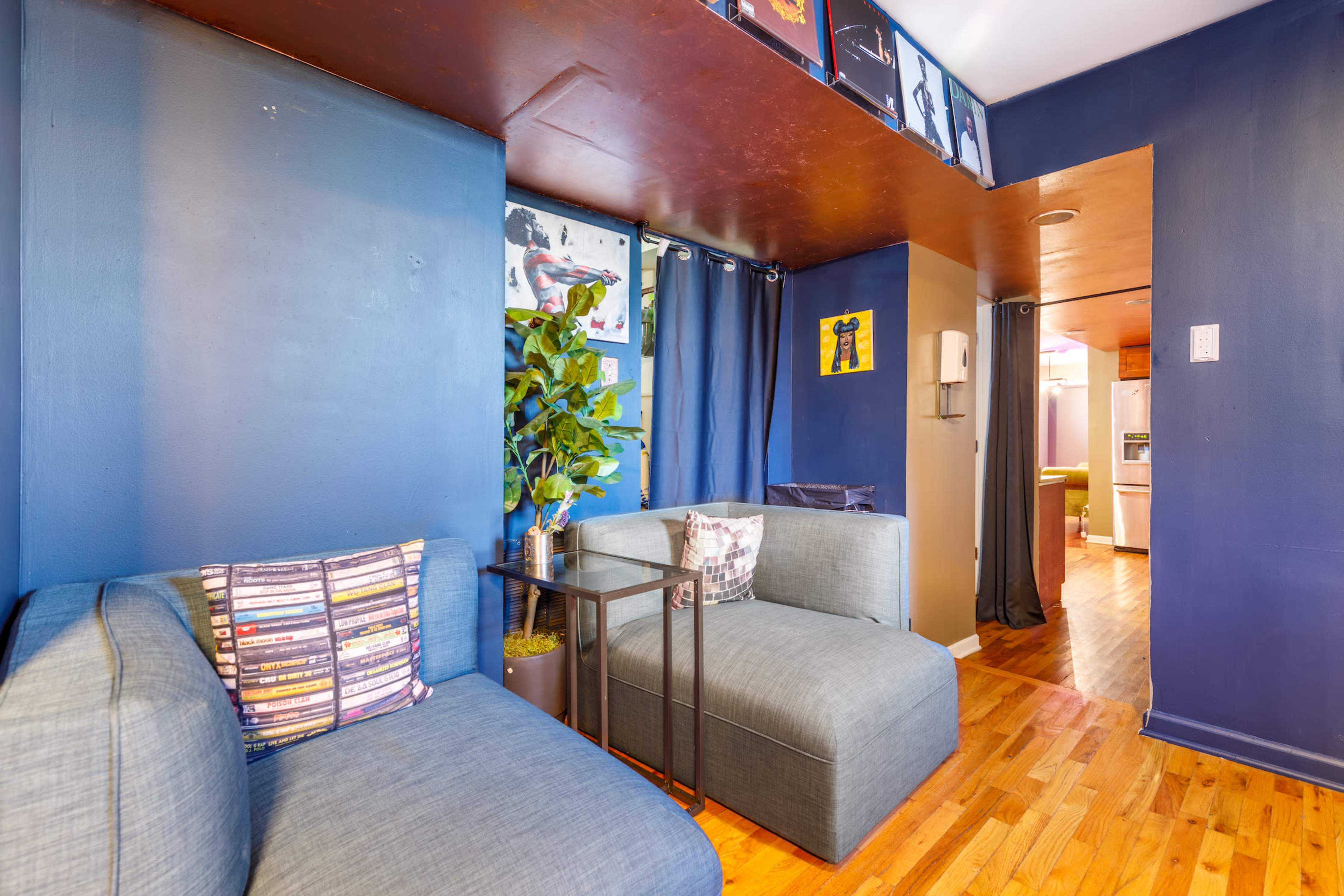 A small living area with blue walls, two gray couches, a side table, and a glimpse of a kitchen in the background.