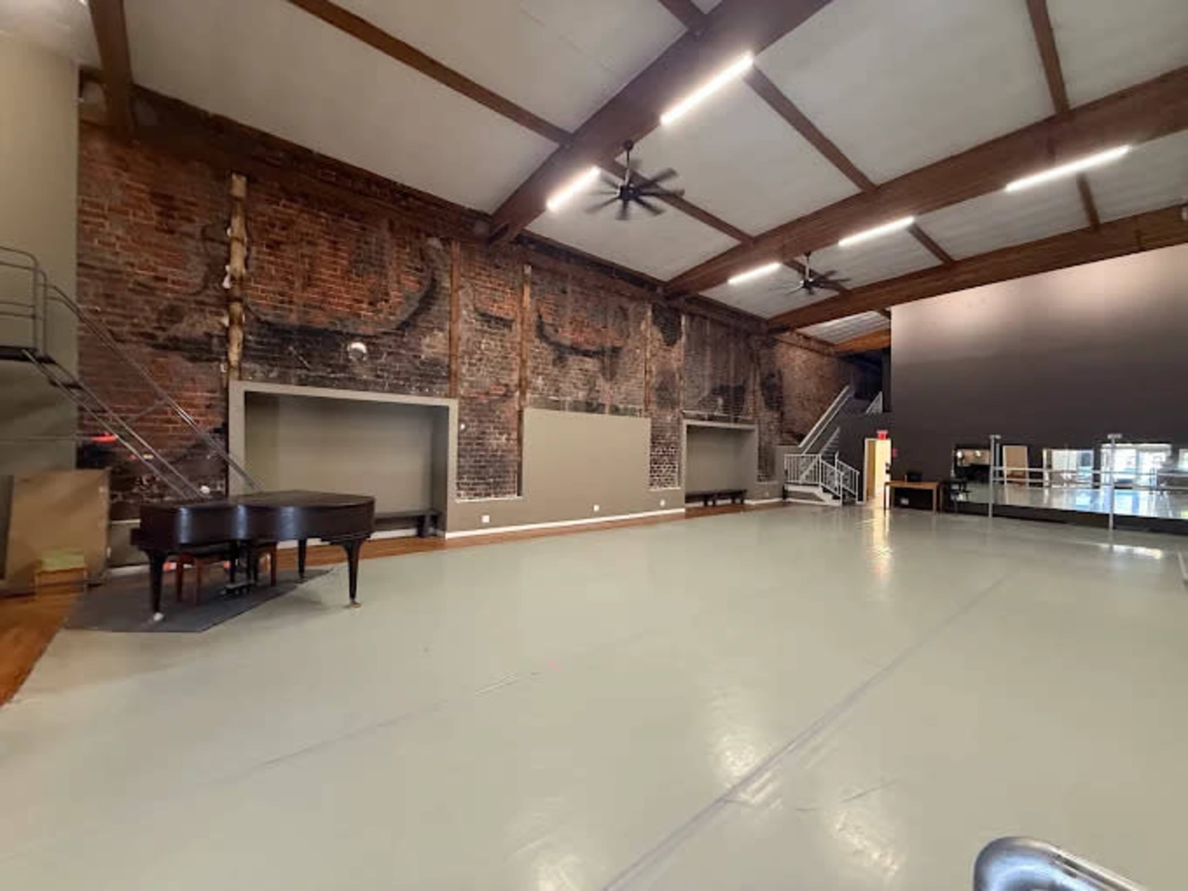 The image shows a spacious interior room with a hardwood floor, exposed brick walls, and a large wall-mounted mirror, accompanied by a grand piano and metal railing leading to an upper level.