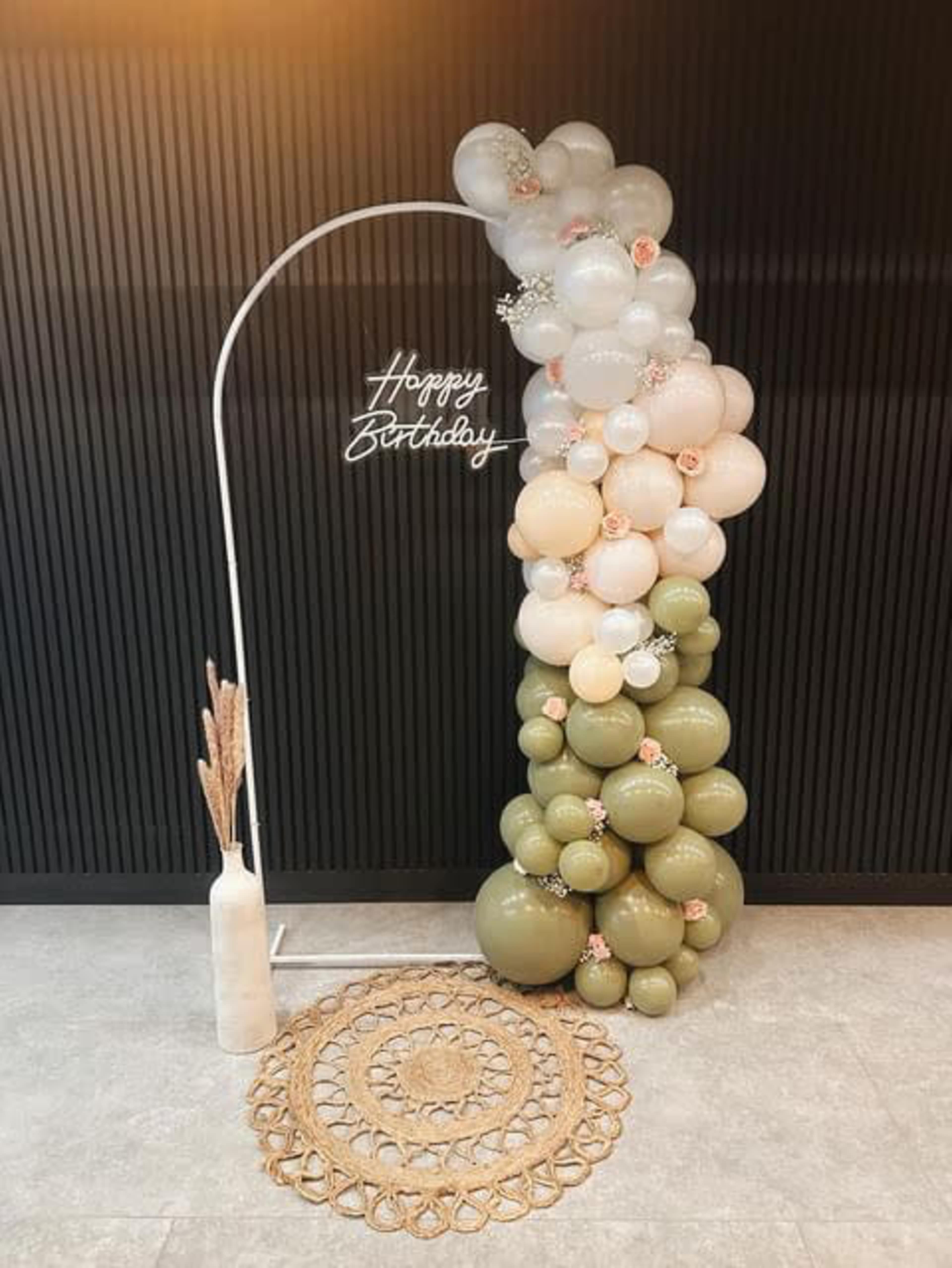 A decorative birthday scene featuring an arch made of assorted balloons in soft colors, with a "Happy Birthday" sign and a woven mat on the floor.