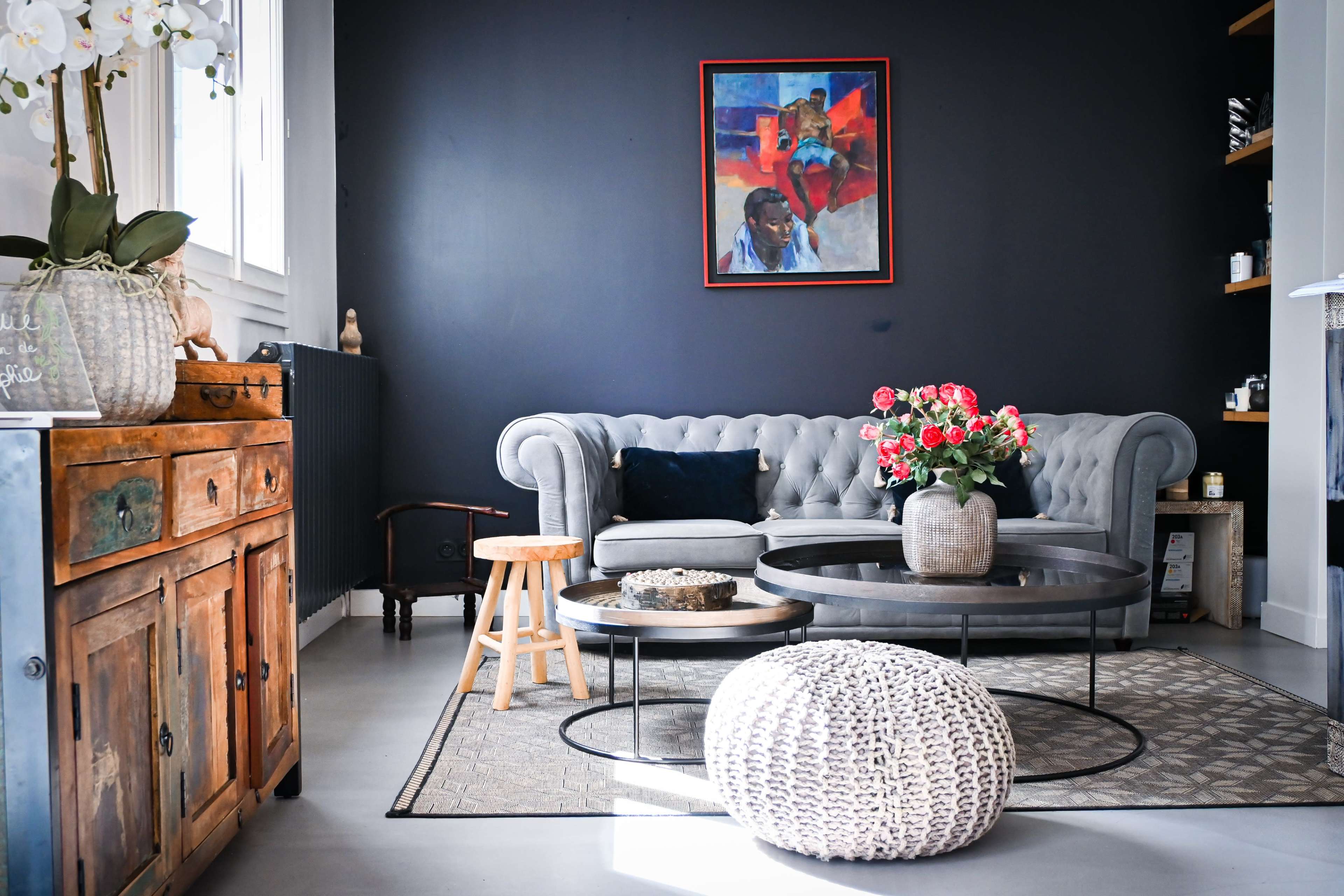 The image shows a stylish living room featuring a grey tufted sofa, a large abstract painting on the wall, and a mix of decorative furniture including a wooden cabinet and various tables.