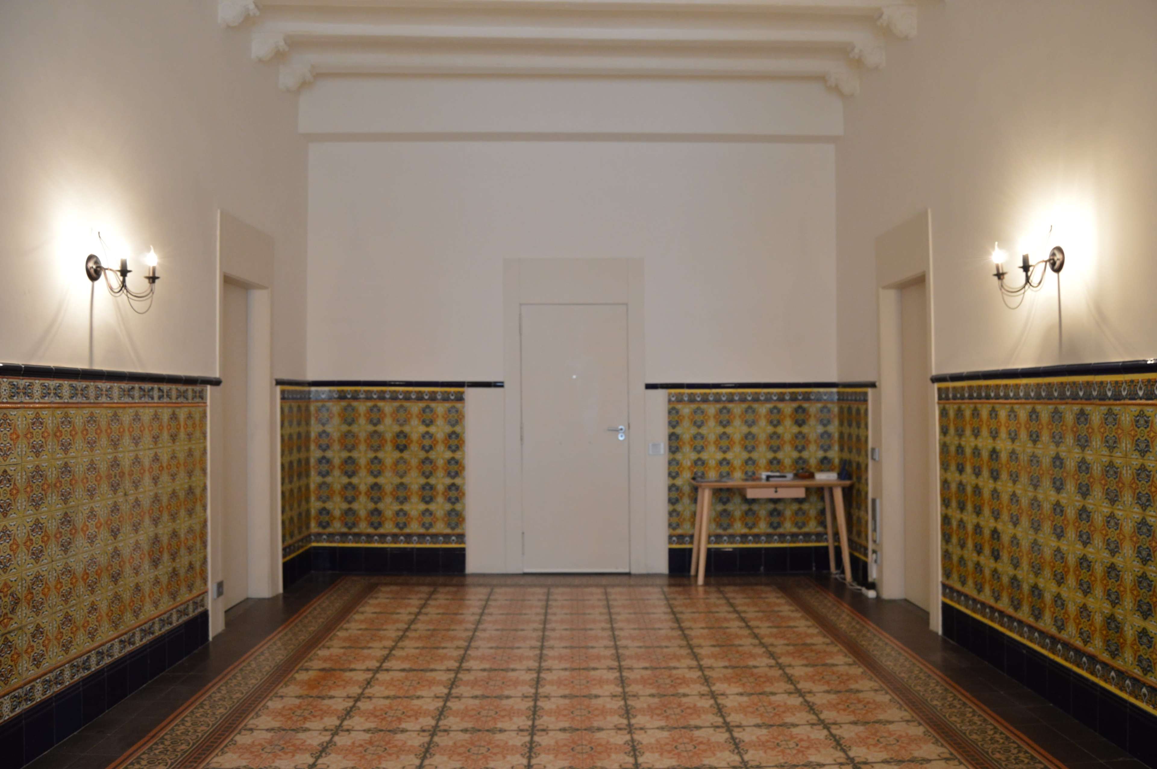 A spacious room with patterned tiled walls and a central tiled floor, featuring two doorways and a table against one wall.
