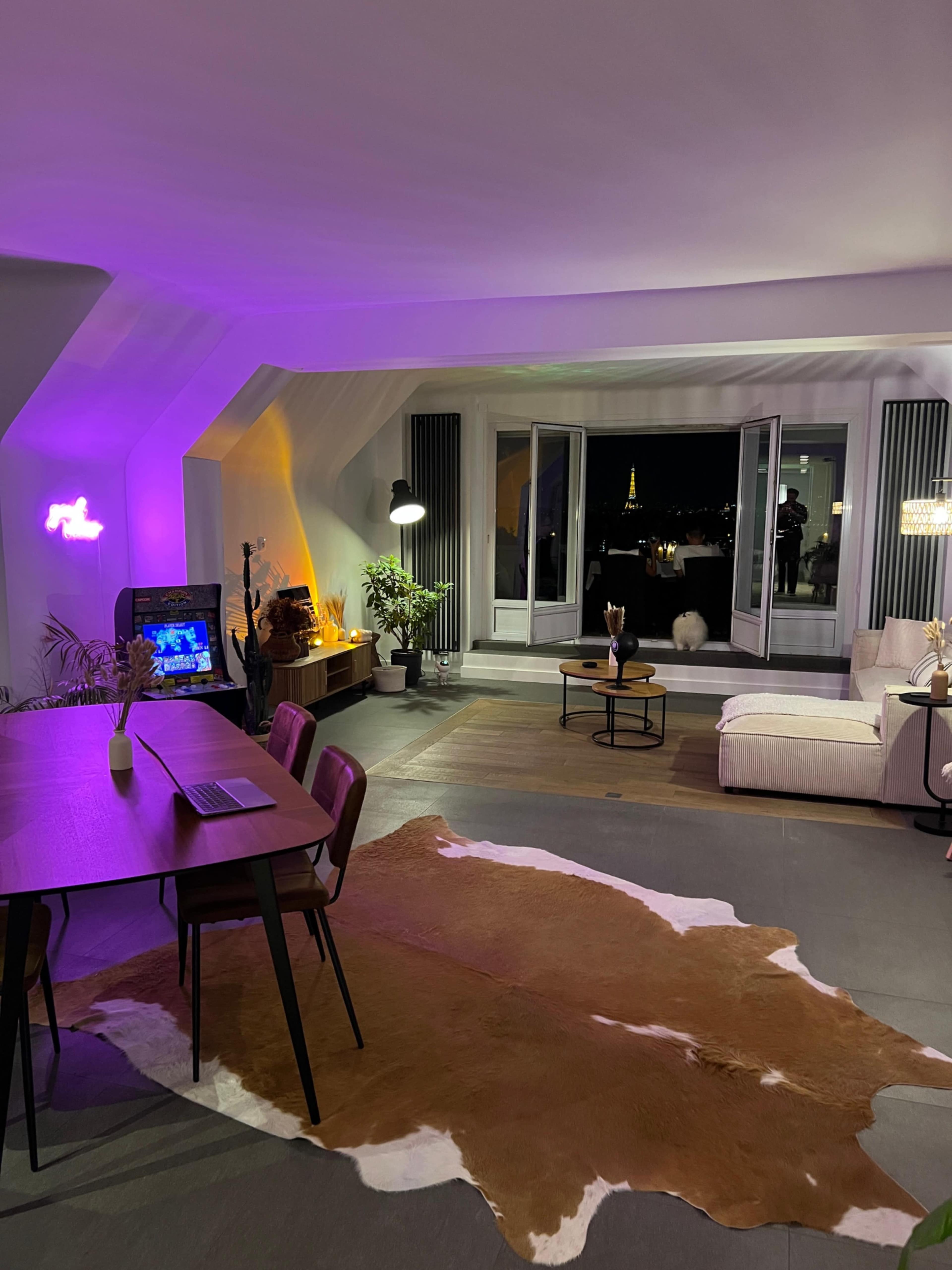 A modern living room features a dining table, a large sofa, and a view of the Eiffel Tower through open windows.