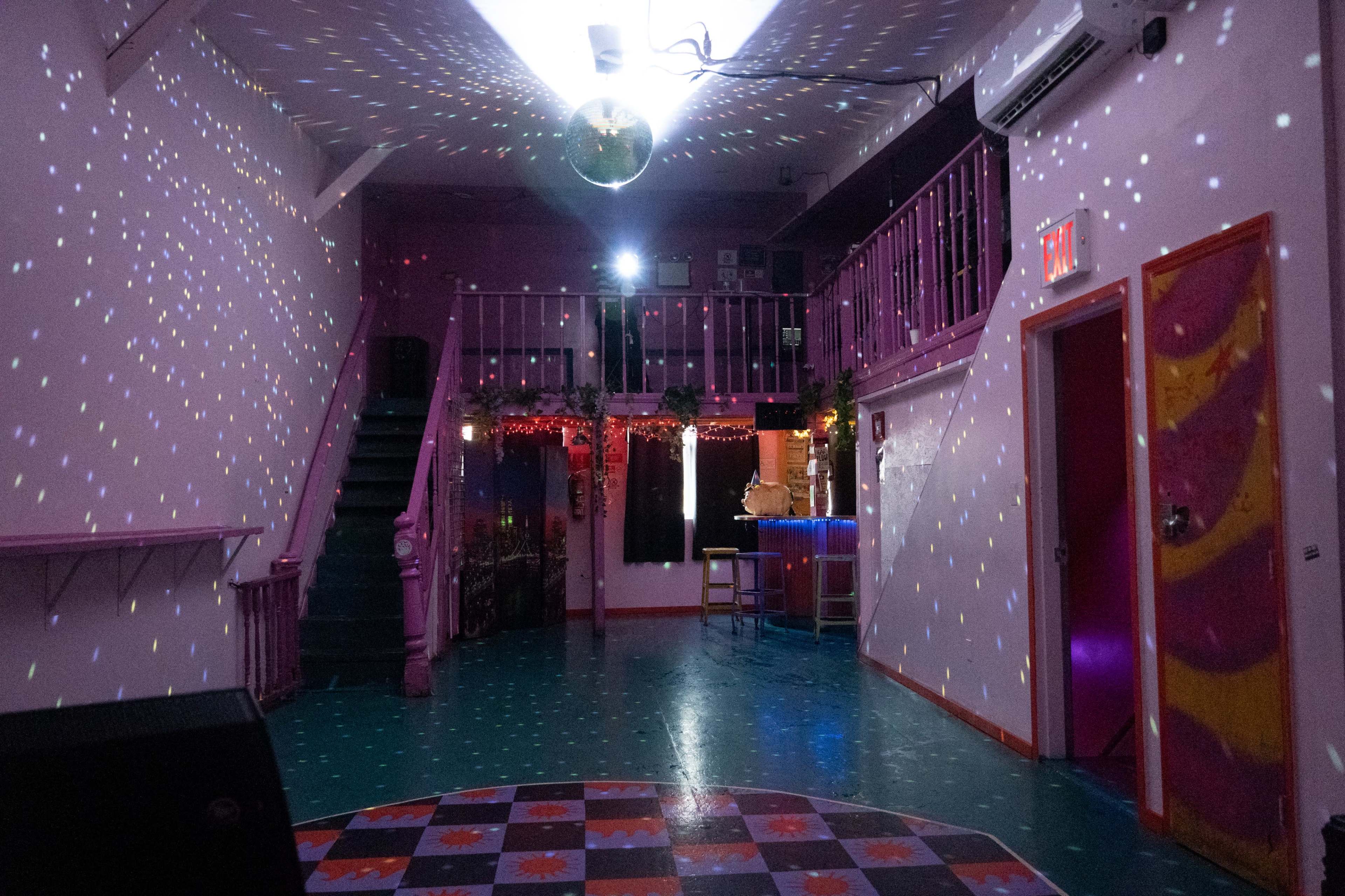 An empty, colorful interior of a venue with a disco ball reflecting lights on the walls and a bar area in the back.